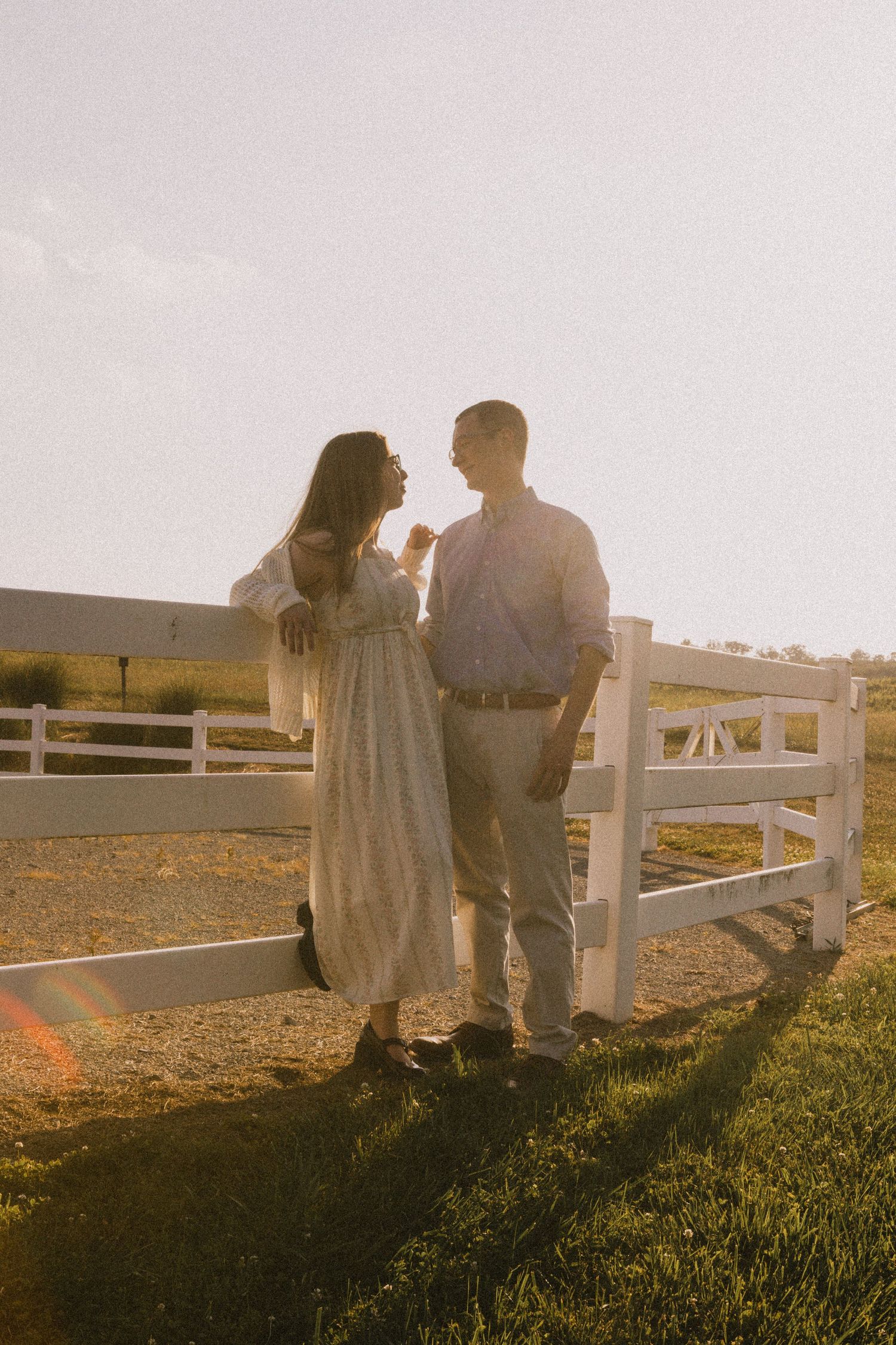 Couple standing together by a white fence at golden hour, sharing a tender moment on a farm.