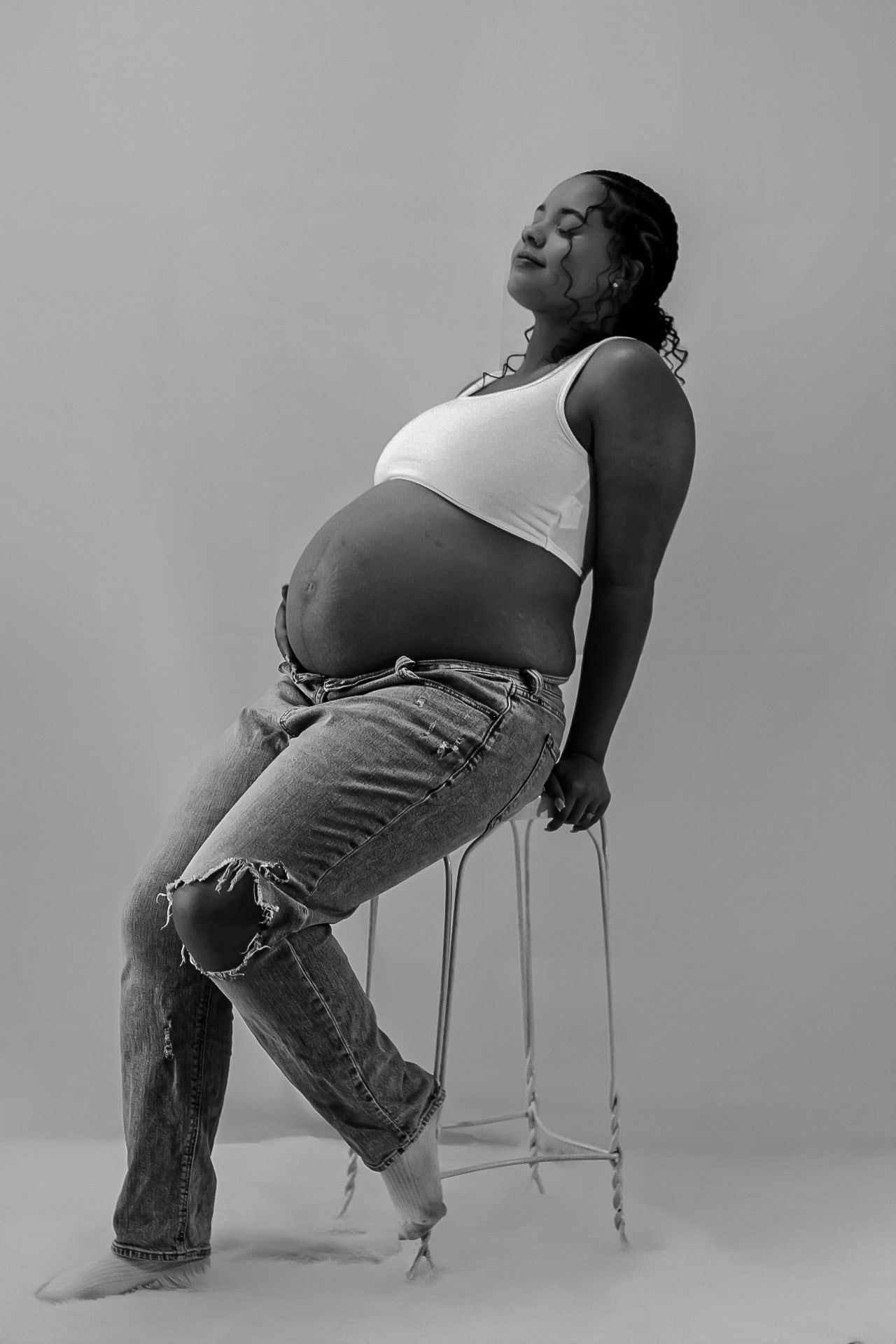 Artistic black and white maternity photo of pregnant person in white tank top and ripped jeans posing on chair.
