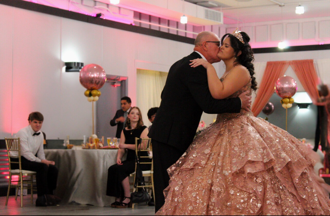 A couple shares their first dance at a quinceañera celebration in a pink-lit ballroom with elegant table settings.