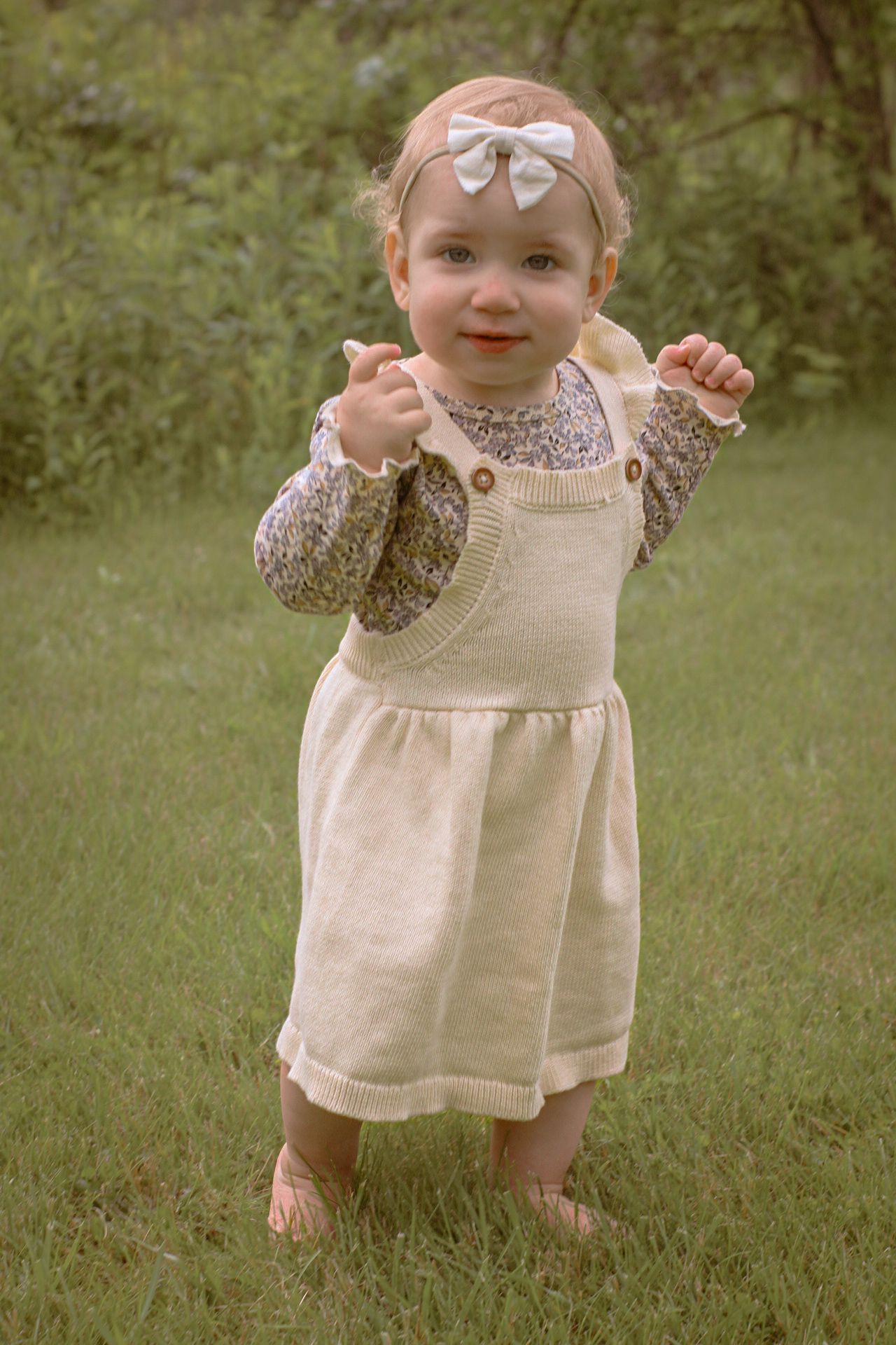 Small child in a vintage-style cream dress and floral accents plays joyfully in a green grassy outdoor setting.