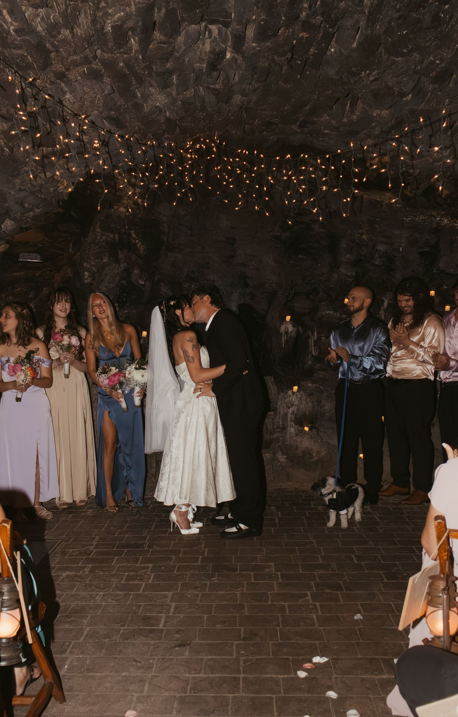 Bride and groom share first kiss in a cave wedding venue with bridesmaids and a dog beside them.