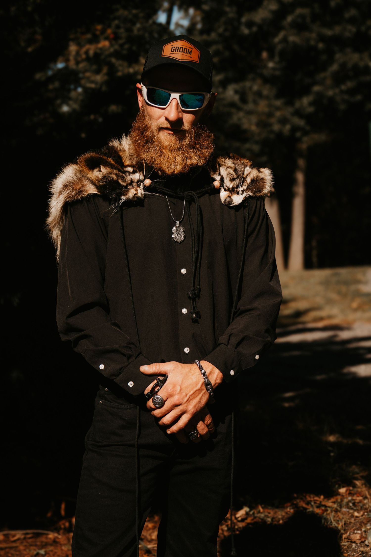 A person in dark clothing stands in dappled sunlight wearing sunglasses and an orange hat against a wooded backdrop.