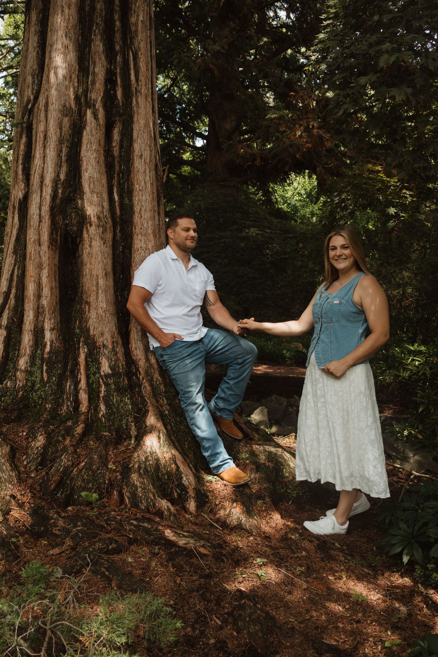 A couple poses by a large redwood tree in a lush forest, man in white shirt and jeans, woman in blue top and white skirt.