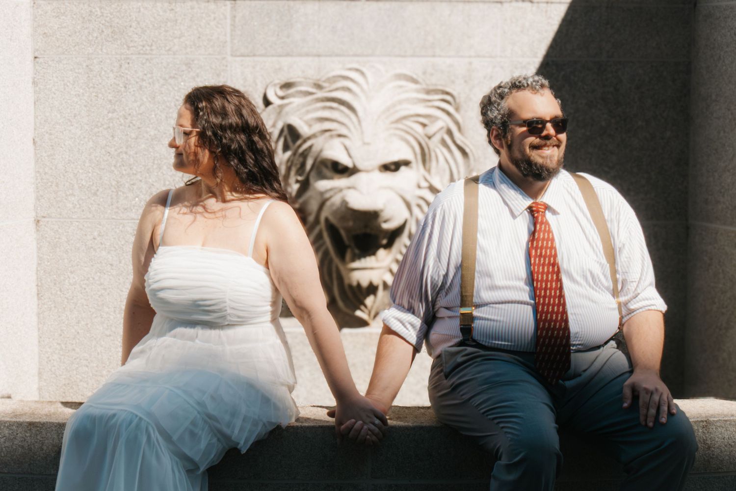 Bride and groom sitting beside a stone lion sculpture, holding hands and smiling on their wedding day.