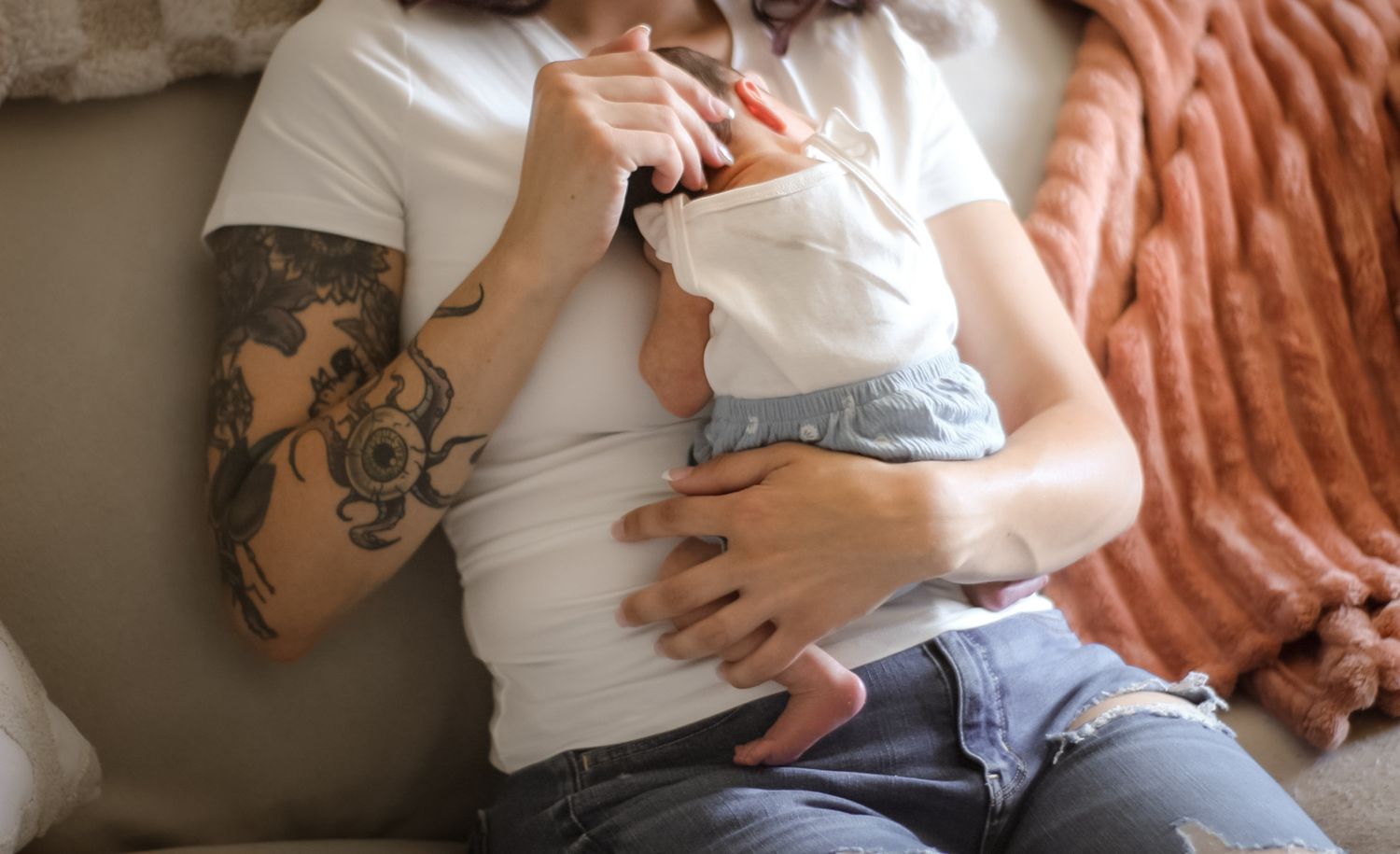 Person with tattoo sleeve holds an infant while sitting on beige couch.