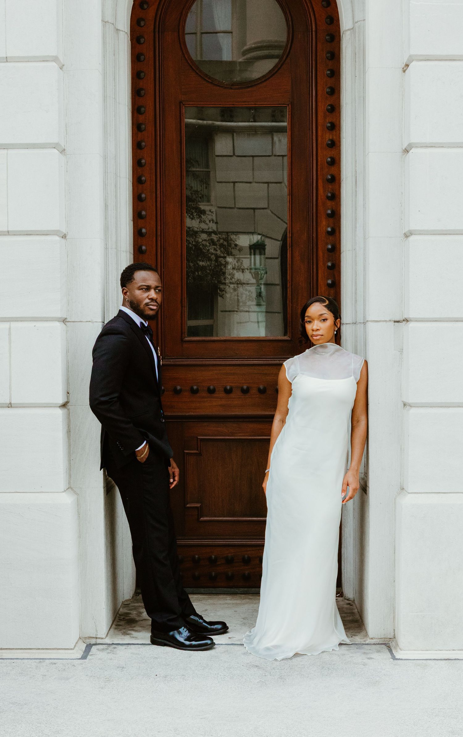 Elegant couple posing before ornate wooden door, groom in black suit, bride in white gown, classic architectural setting.