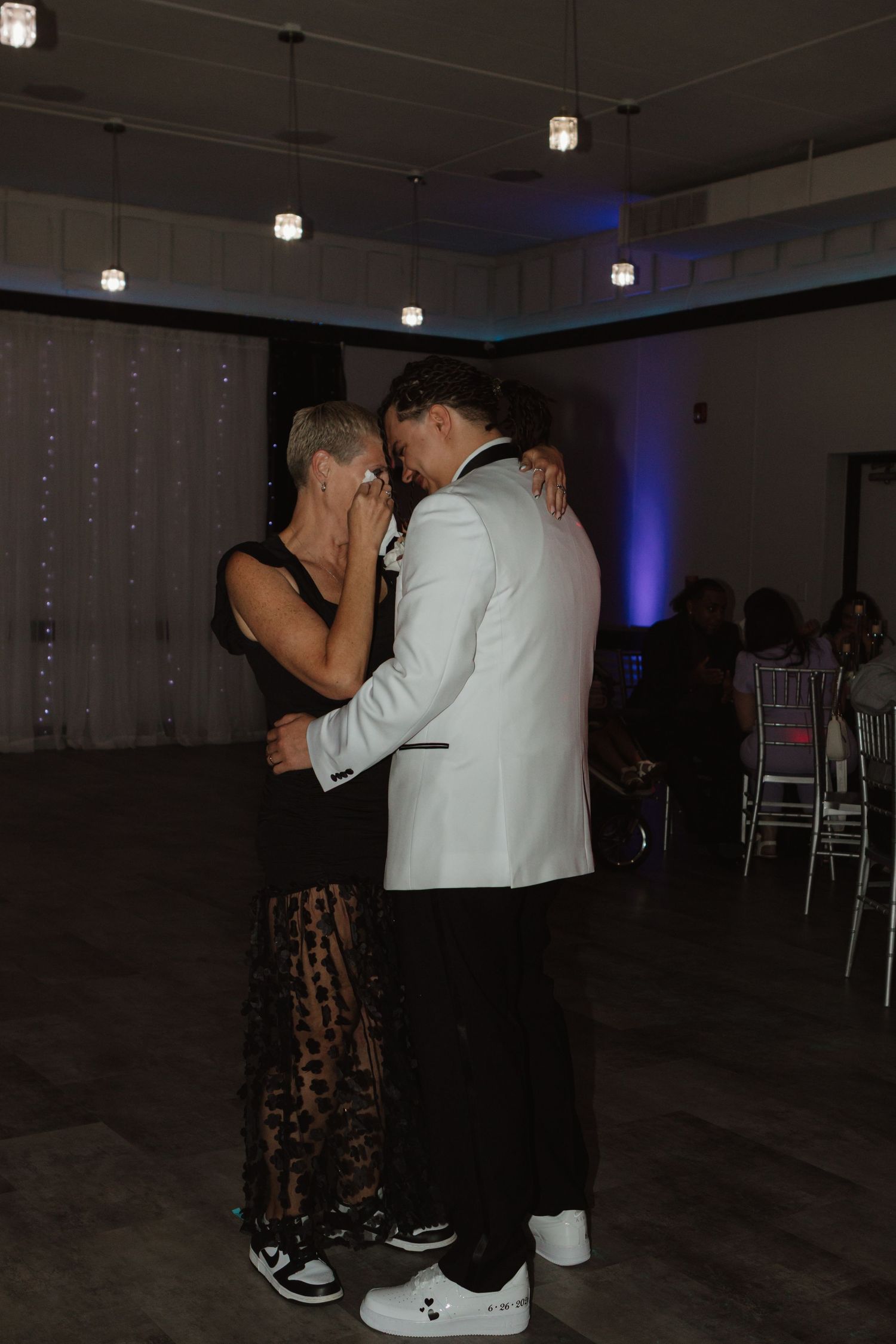 A young man in a white tuxedo jacket shares an emotional slow dance with a woman in a black lace dress at a formal event.