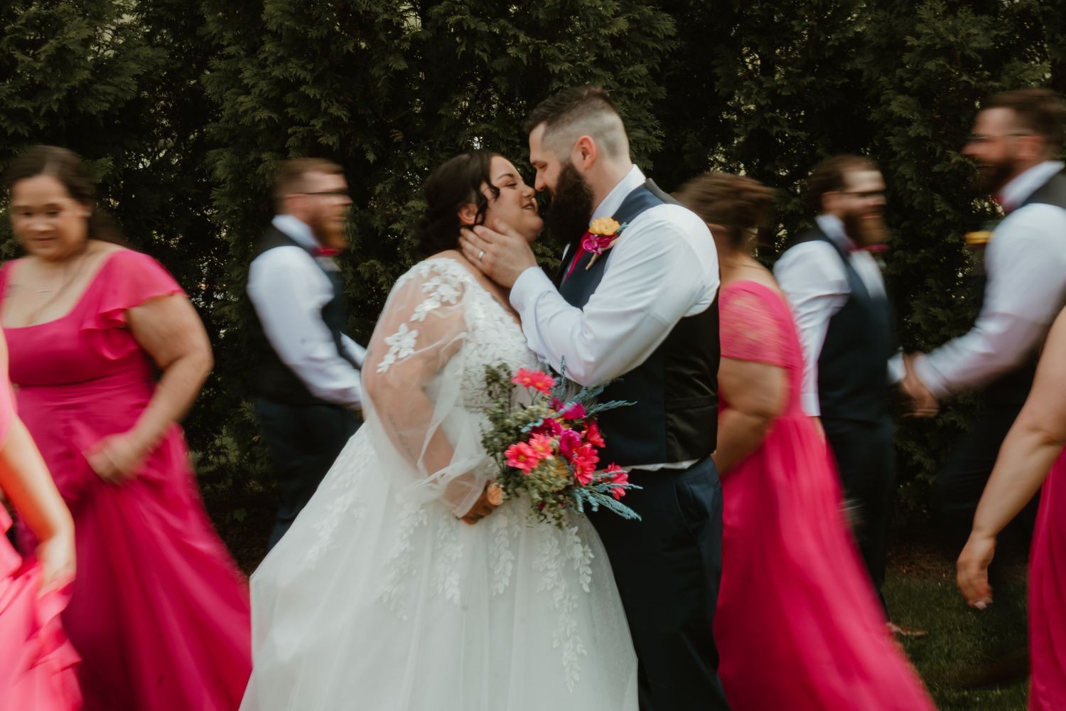 Bride and groom share a kiss while bridesmaids in pink dresses and groomsmen walk past in a blurred motion outdoor wedding.