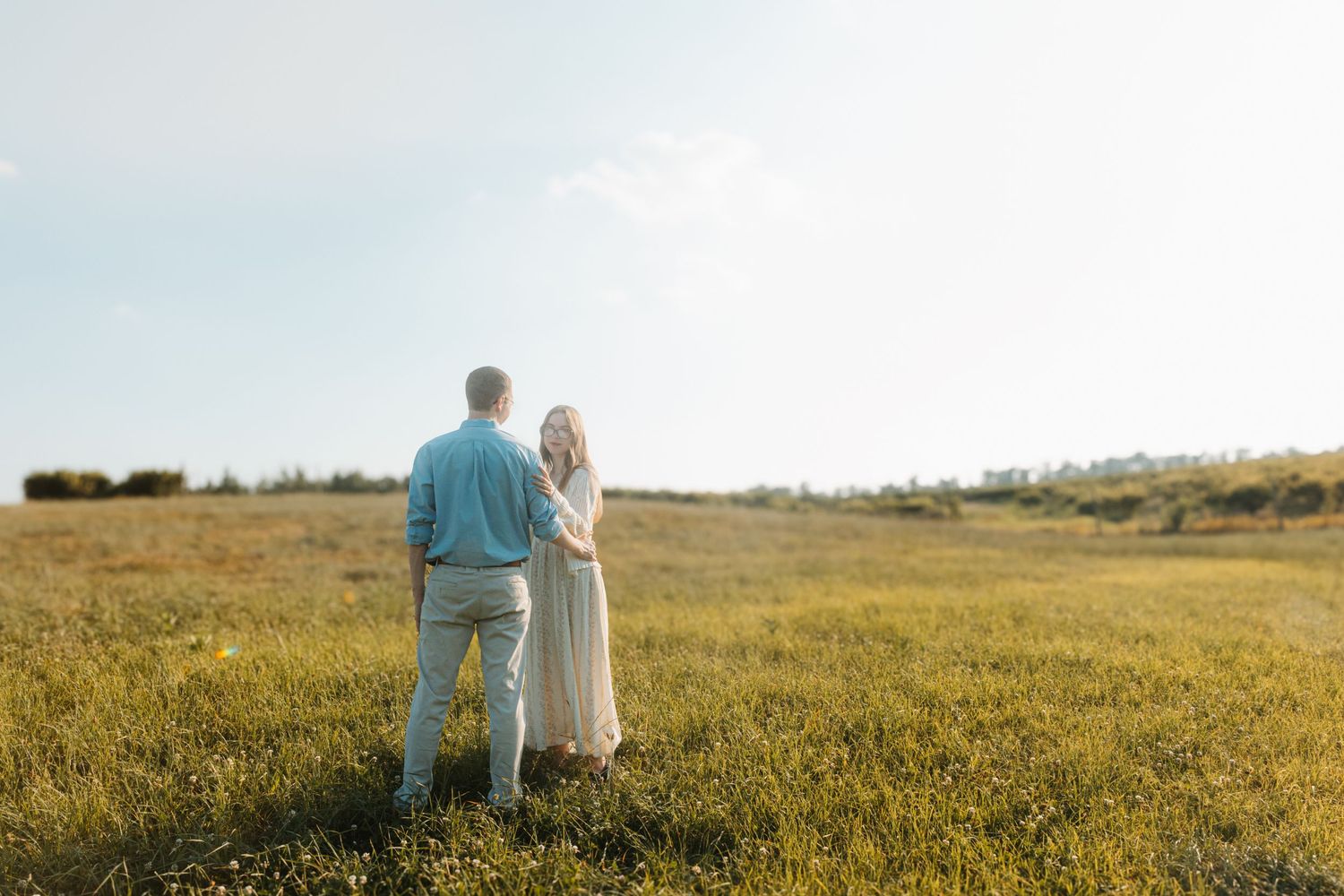 Couple holding hands while standing in a vast open meadow under a clear sky viewed from behind.