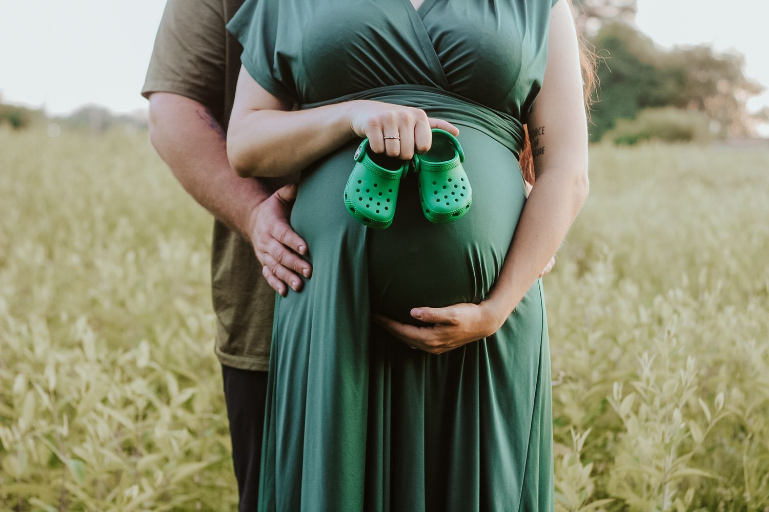 Close-up of a pregnant belly with tiny green baby shoes held against it in a sunny field.