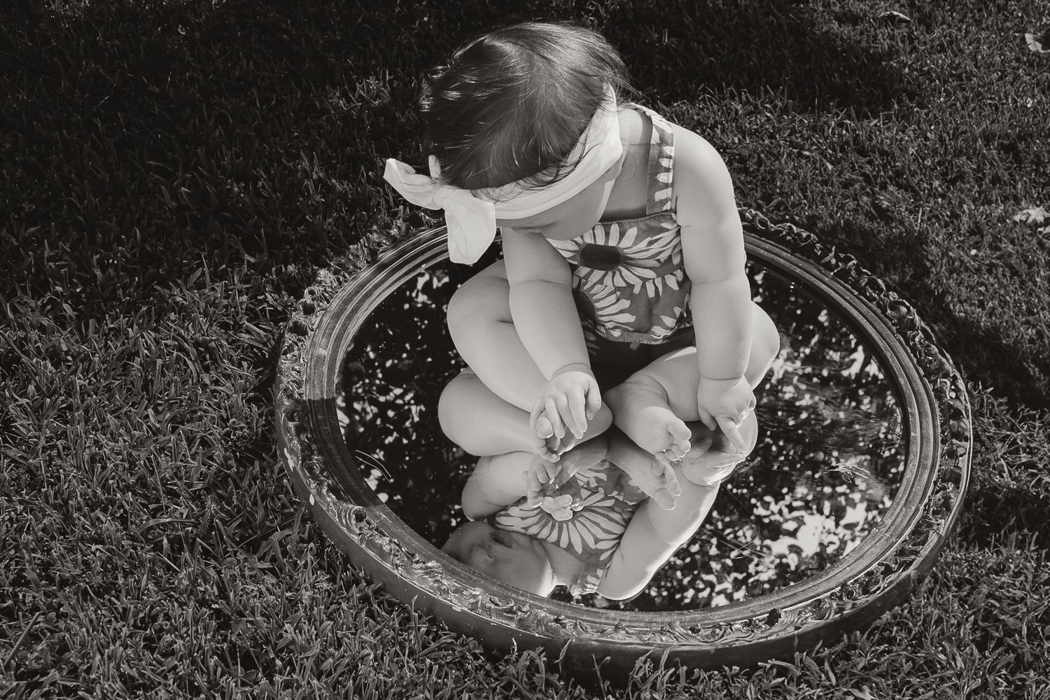 Vintage black and white photo of a child sitting on an ornate mirror outdoors.