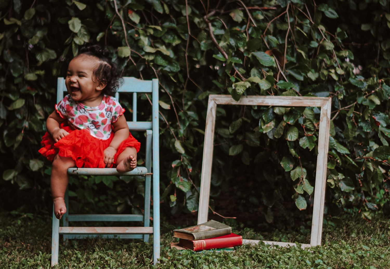 Baby in red dress with floral collar sitting on turquoise chair against green foliage background.