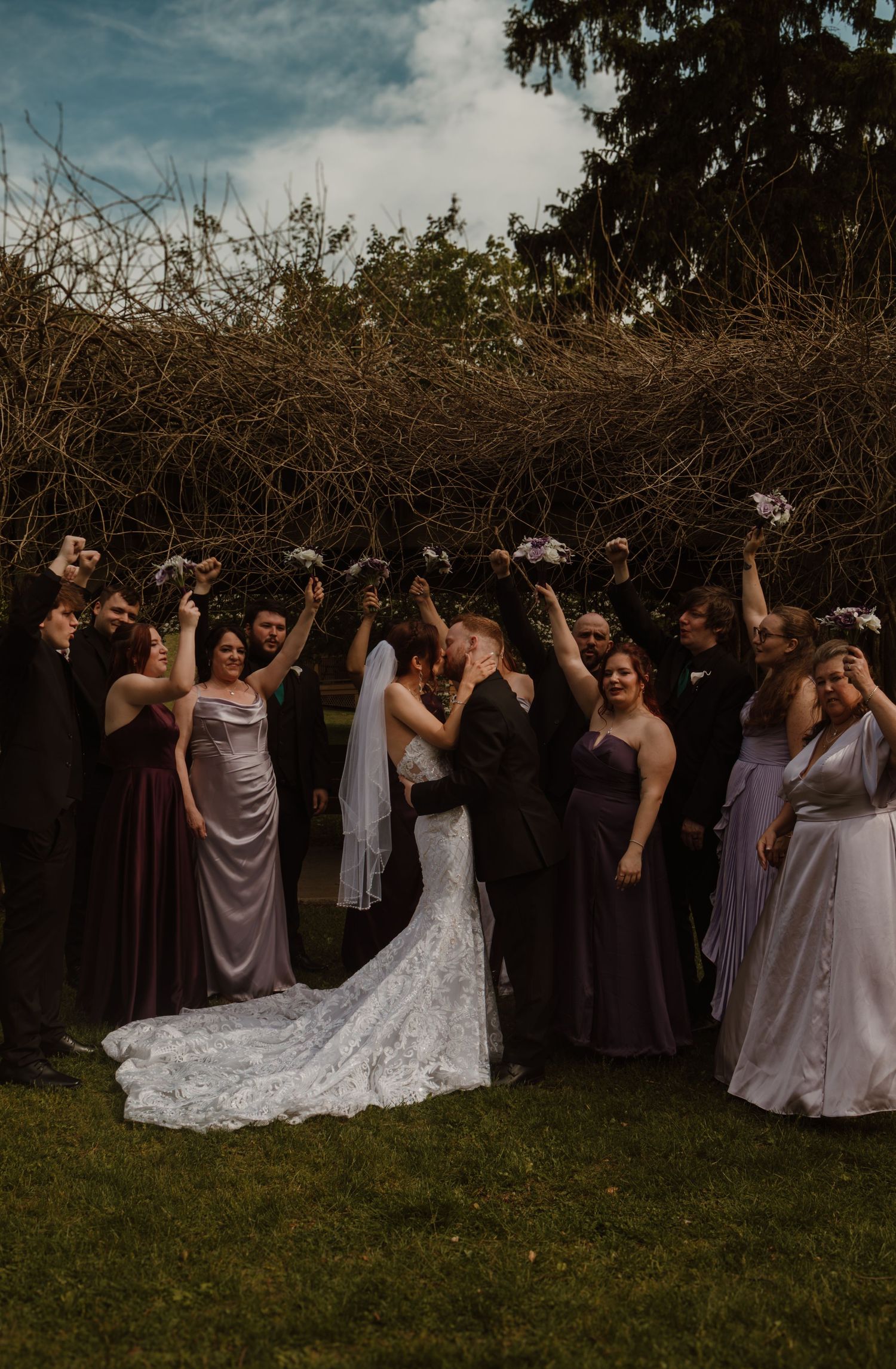 Wedding party celebrates outdoors as bride and groom share a kiss surrounded by bridesmaids in purple dresses.