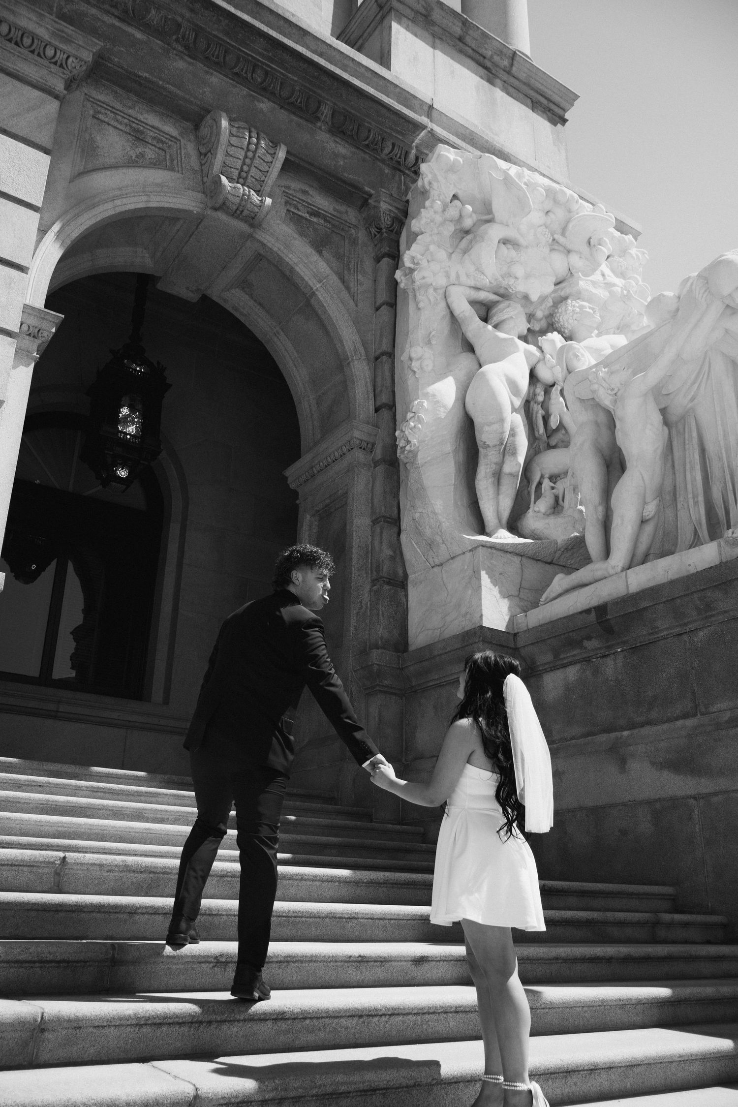 Black and white photo of a couple holding hands on grand stone steps outside a classical building with ornate sculptures.