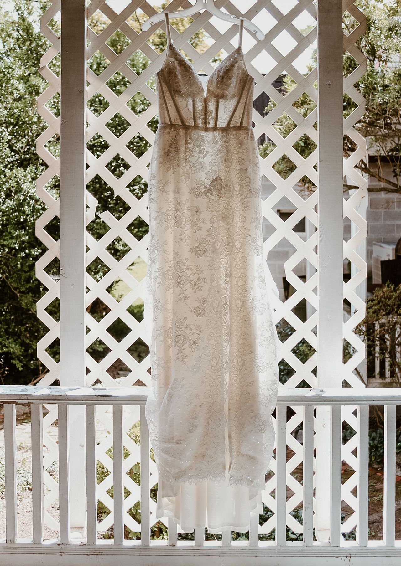 A delicate lace wedding dress hangs against white lattice backdrop in natural outdoor lighting.