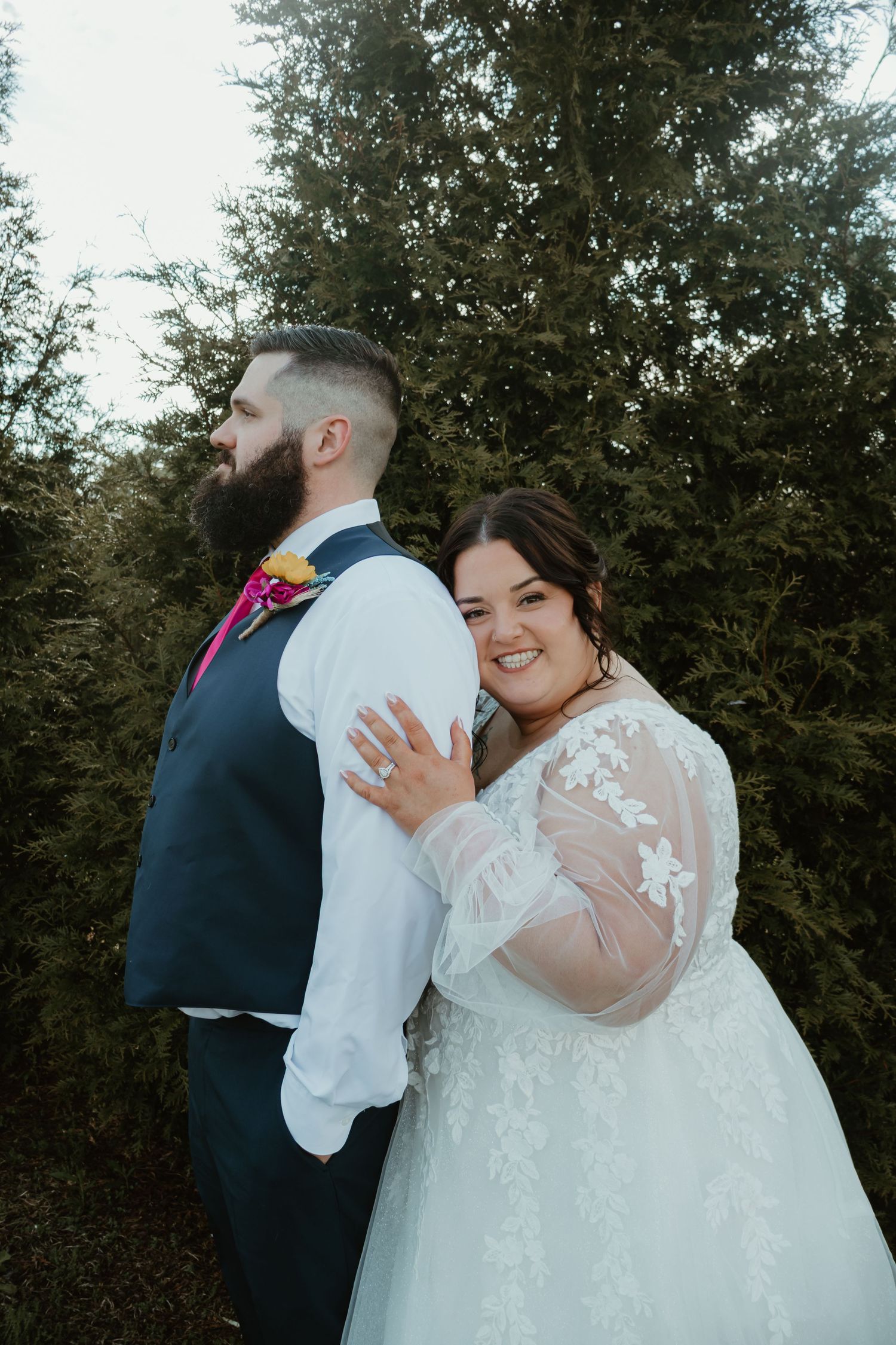 Bride in lace gown embraces groom in navy vest among evergreen trees at outdoor wedding.
