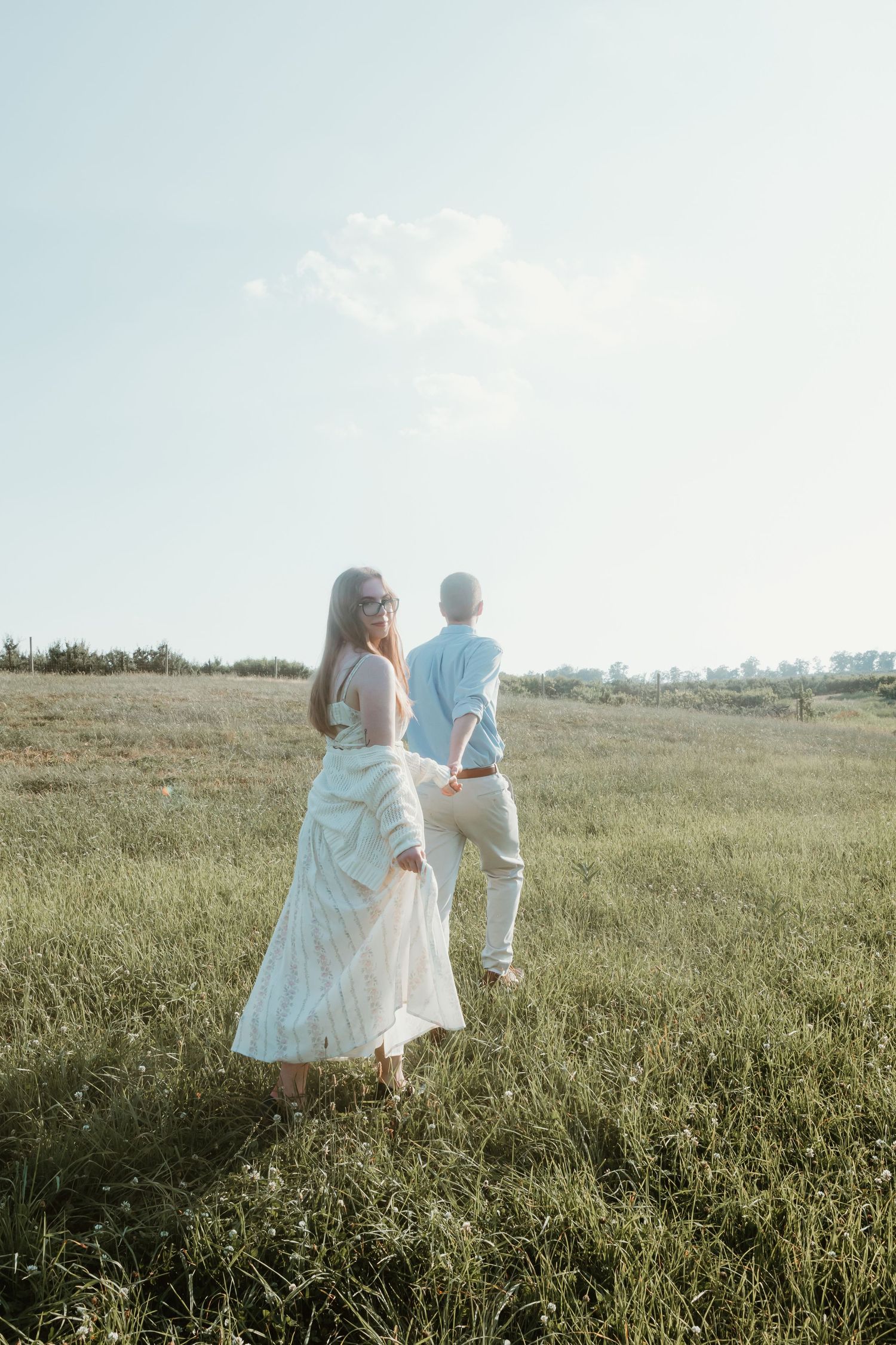 Woman in white dress holding hands with man in light blue shirt walking through green field.