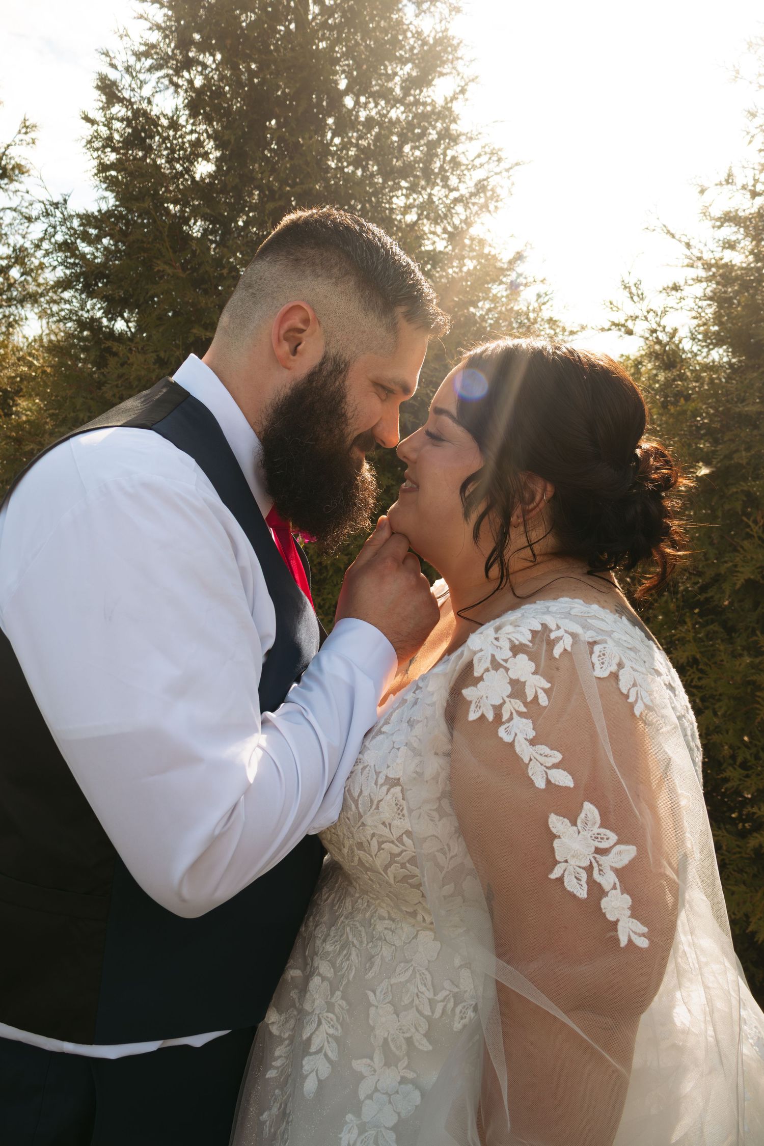 Bearded groom in vest tenderly holds brides chin as they share a romantic moment in golden sunlight wearing wedding attire.