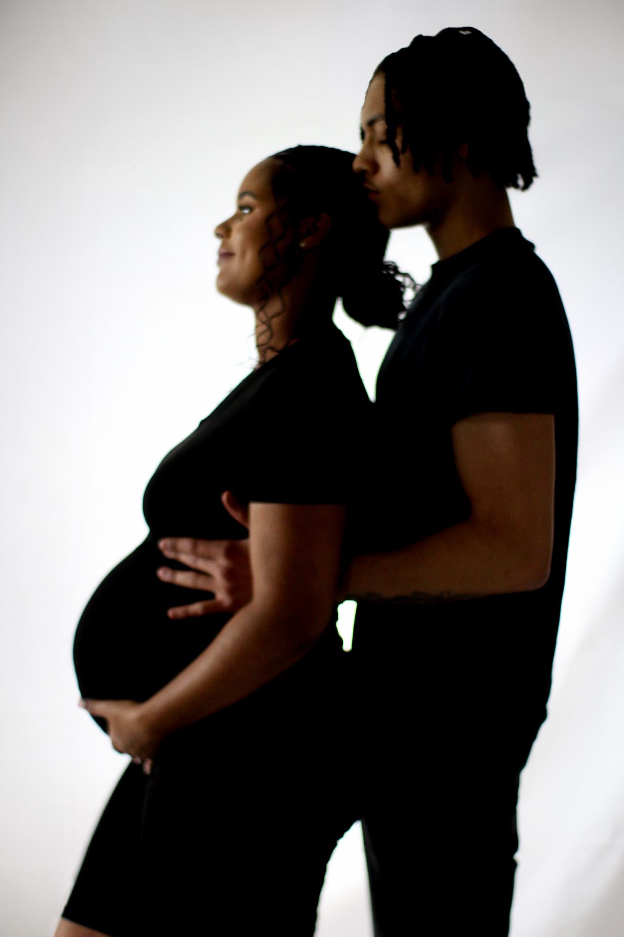 Side-lit portrait of expecting couple in black clothing embracing.