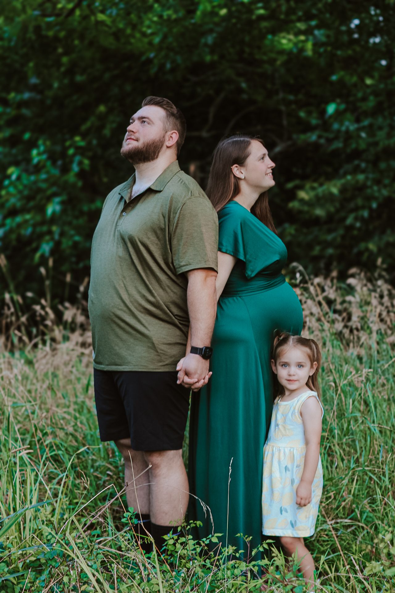 A family of three stands together in a field, with the woman wearing a teal maternity dress.