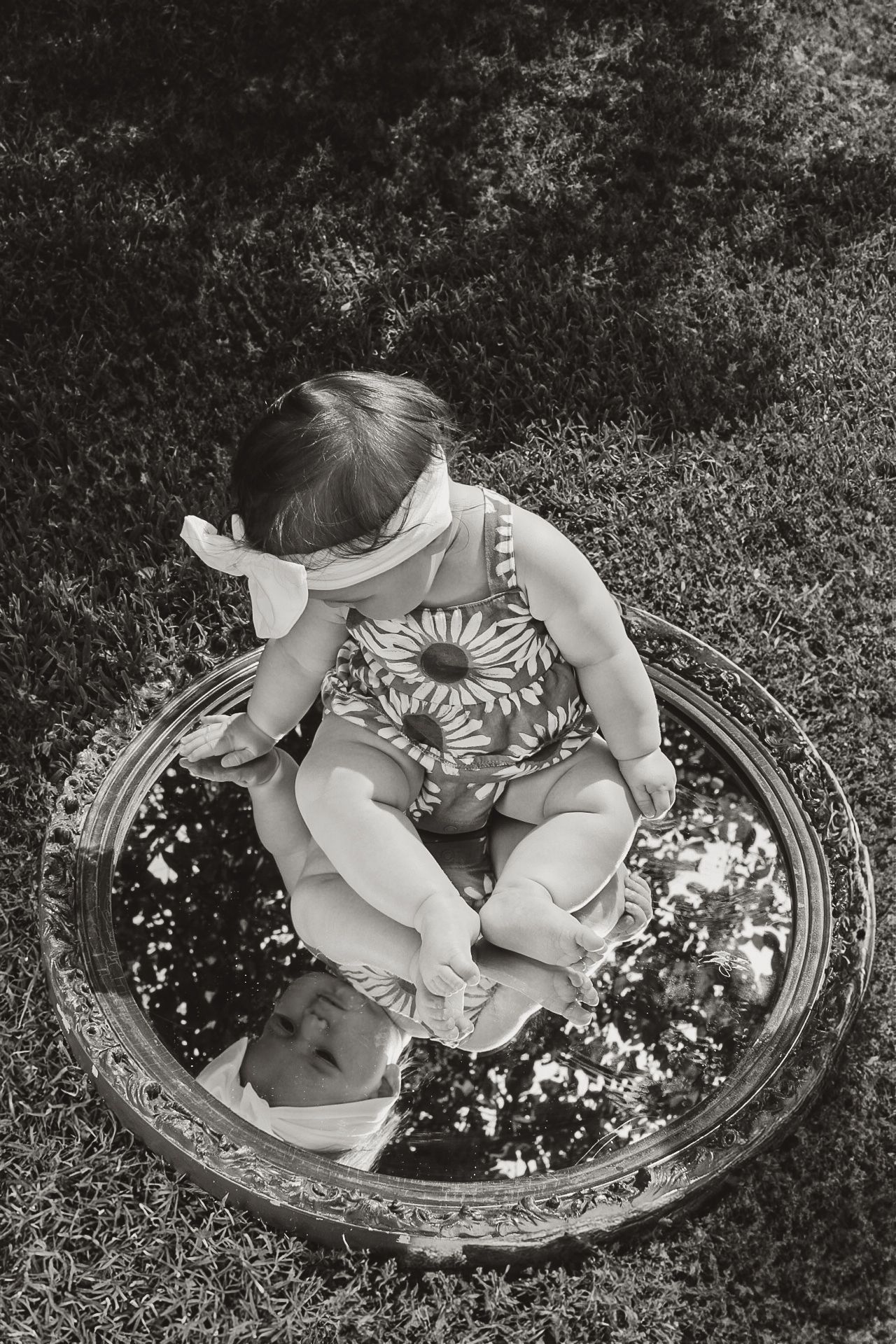 Black and white artistic photo of a child sitting on a vintage mirror in a garden.
