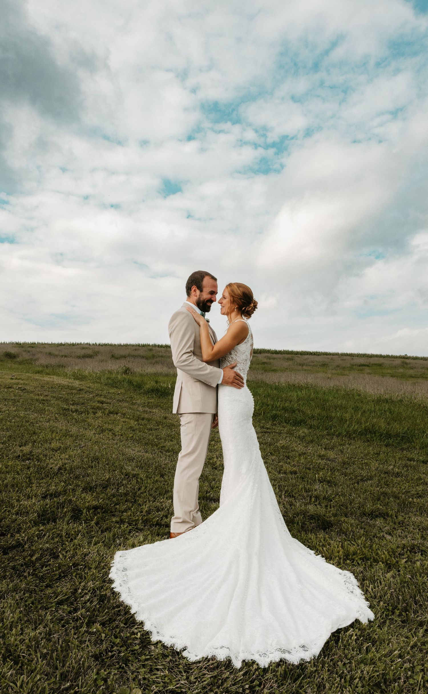 A bride in a white gown with a long train stands with a groom in a tan suit in a green field under a cloudy sky.