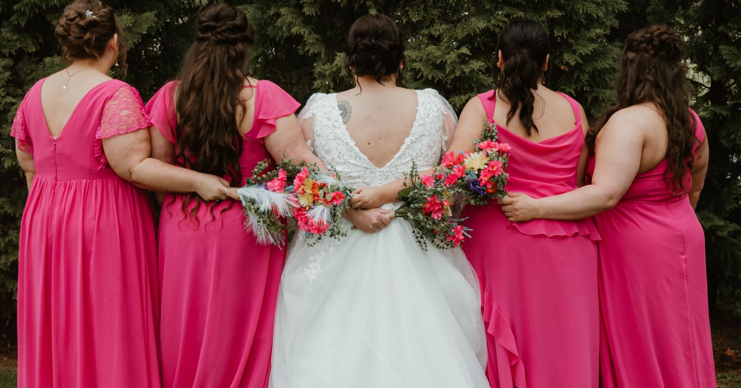 Bride in white lace gown stands with four bridesmaids in pink dresses holding colorful bouquets, viewed from behind.