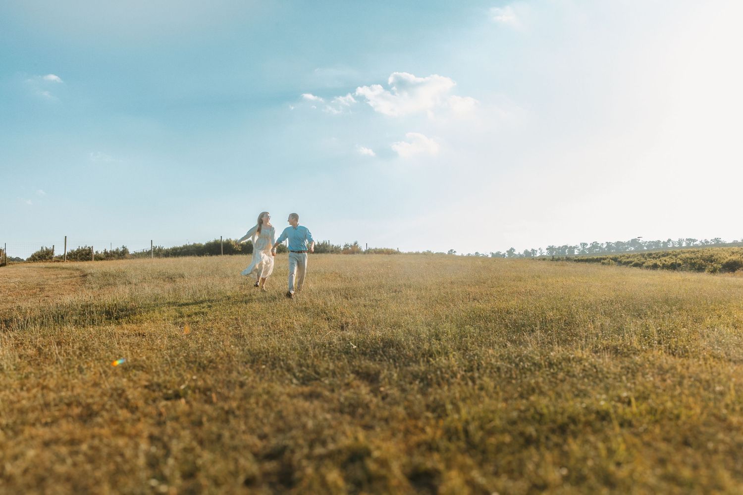 Couple holding hands running through a sunny open meadow under a bright blue sky.
