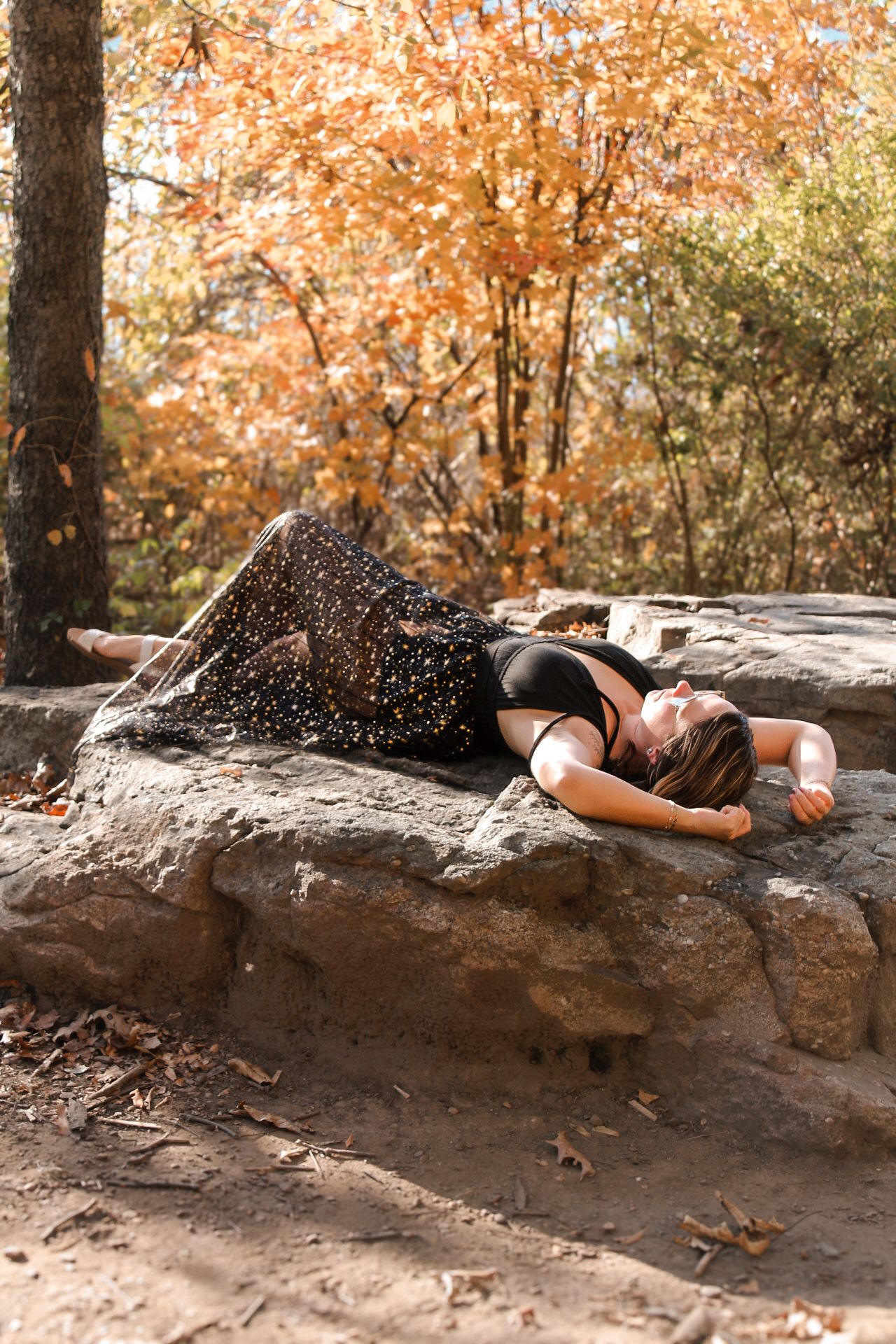 A person in a black dress lying on a rock formation surrounded by autumn foliage in a natural outdoor setting.