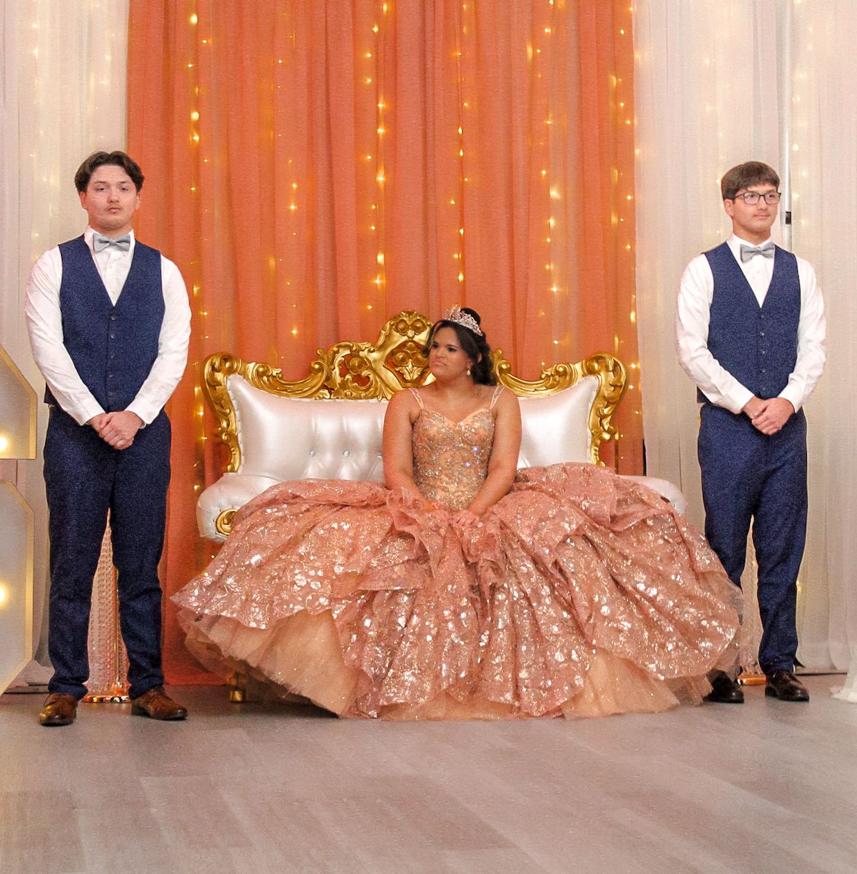 A quinceañera celebrant in rose gold gown seated between two attendants in navy vests.