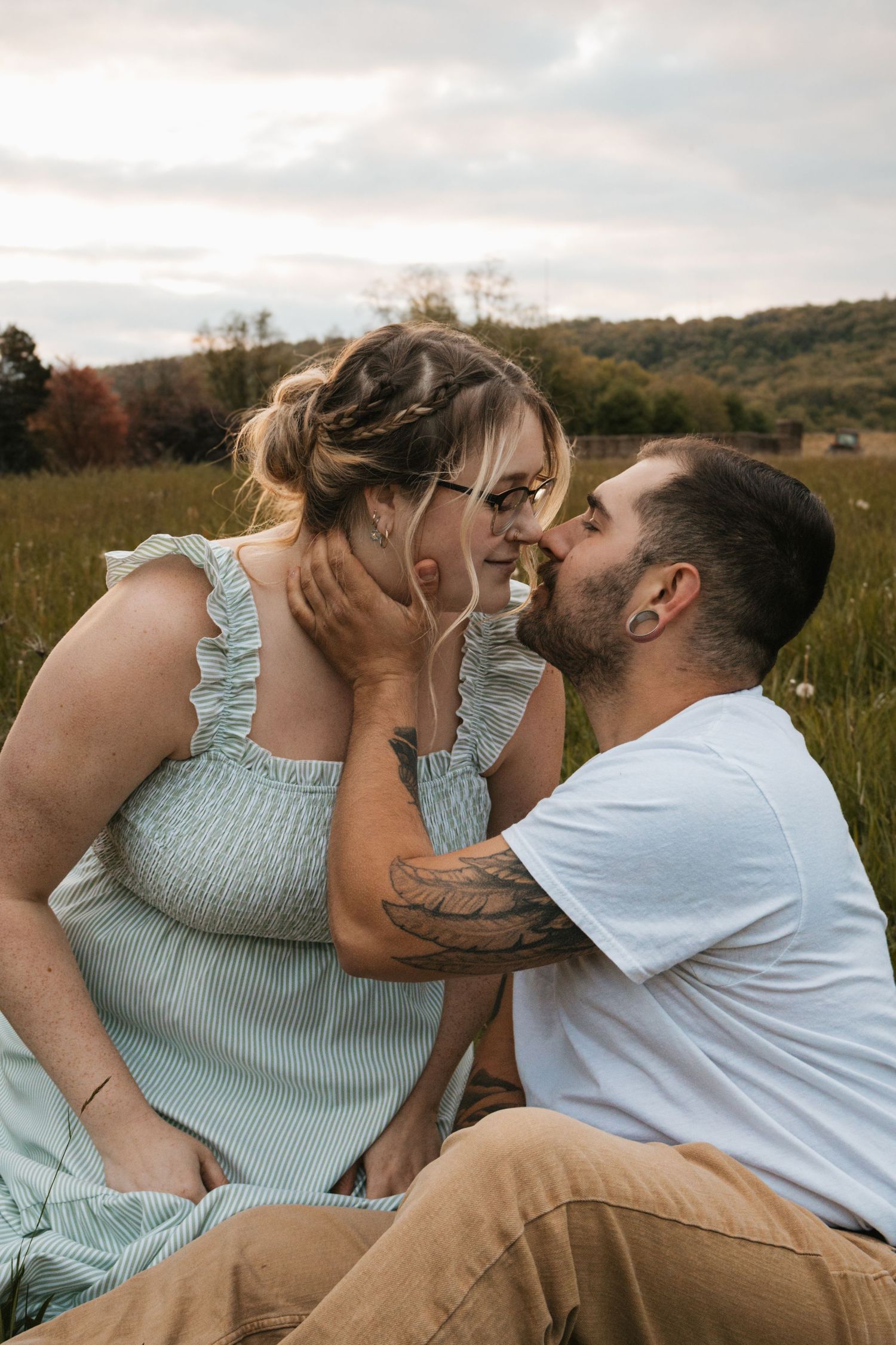Couple sharing a romantic kiss while sitting in a lush green field at sunset, woman in mint dress, man in white tee.