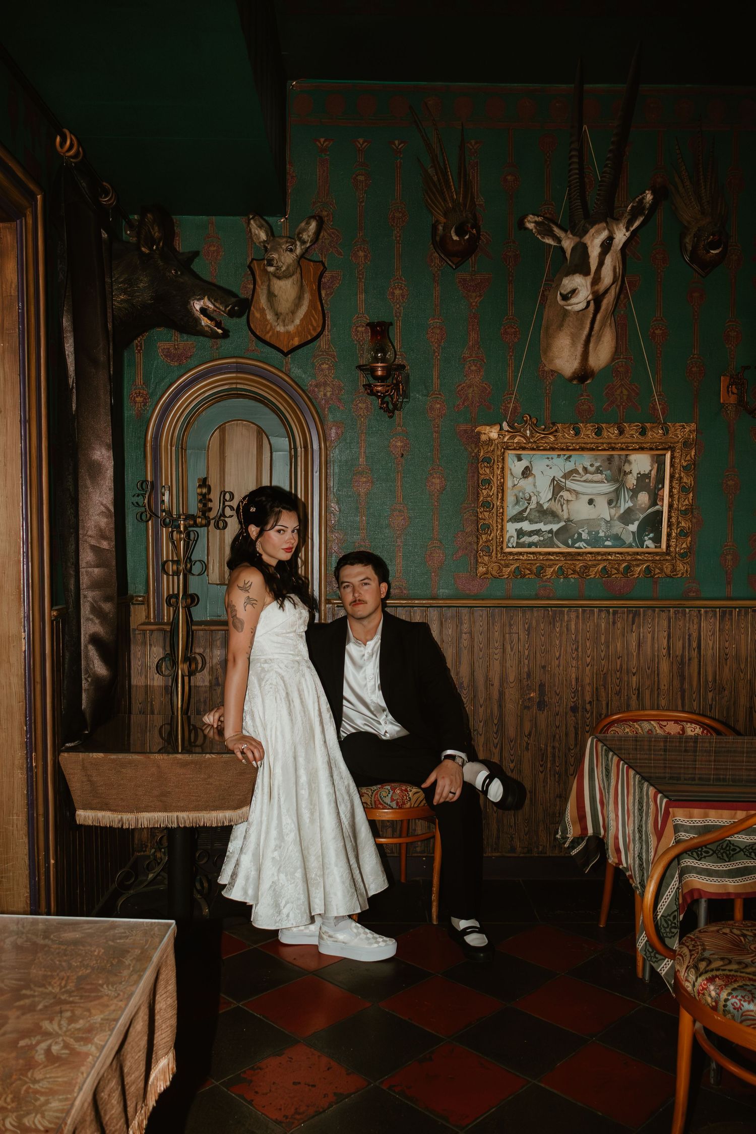 Bride in white dress and groom in black suit pose in a vintage room with taxidermy mounts and green wallpaper.