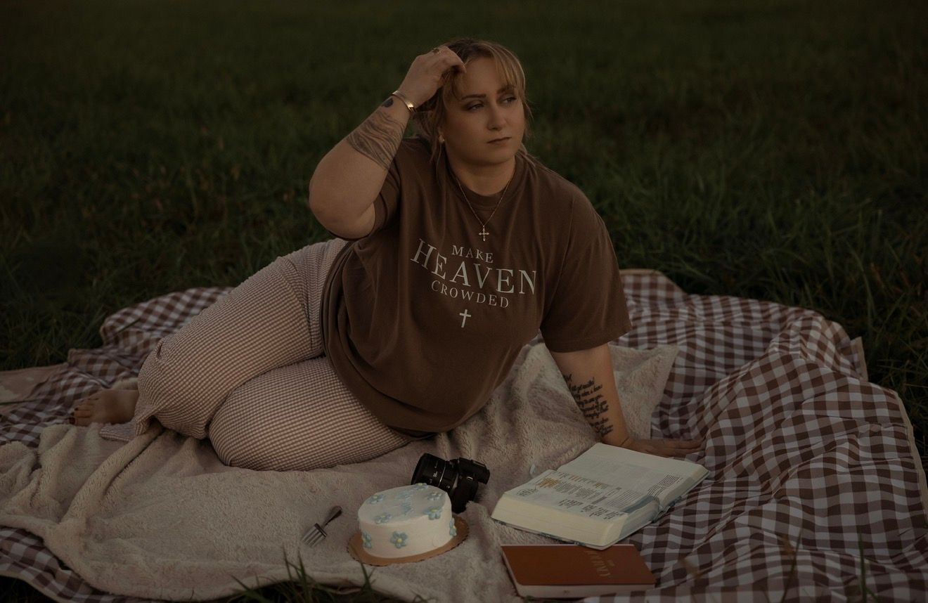 Woman in brown graphic tee sits on plaid blanket in grass field at dusk with books and candle.