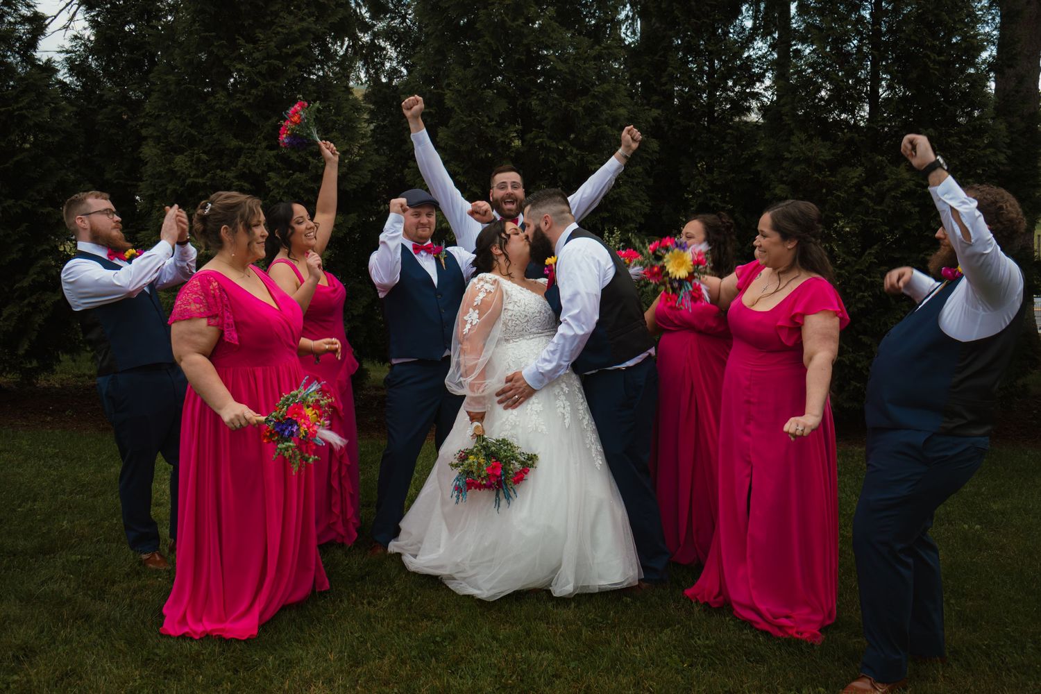 Wedding party celebrating outdoors, bridesmaids in red dresses cheering as bride and groom share a kiss.