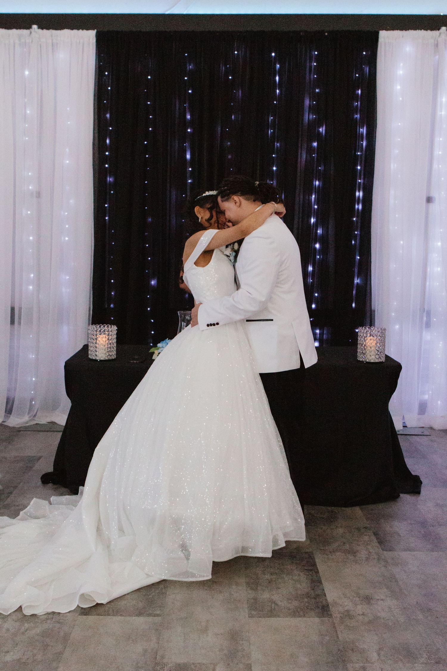 Bride and groom share a romantic kiss on the dance floor in front of a black and white curtain backdrop with fairy lights.