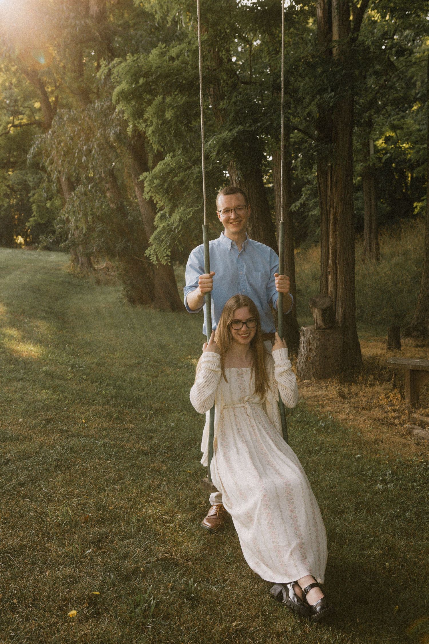 A couple enjoys a sunny garden swing, woman in white dress and man in blue shirt, surrounded by lush green trees.