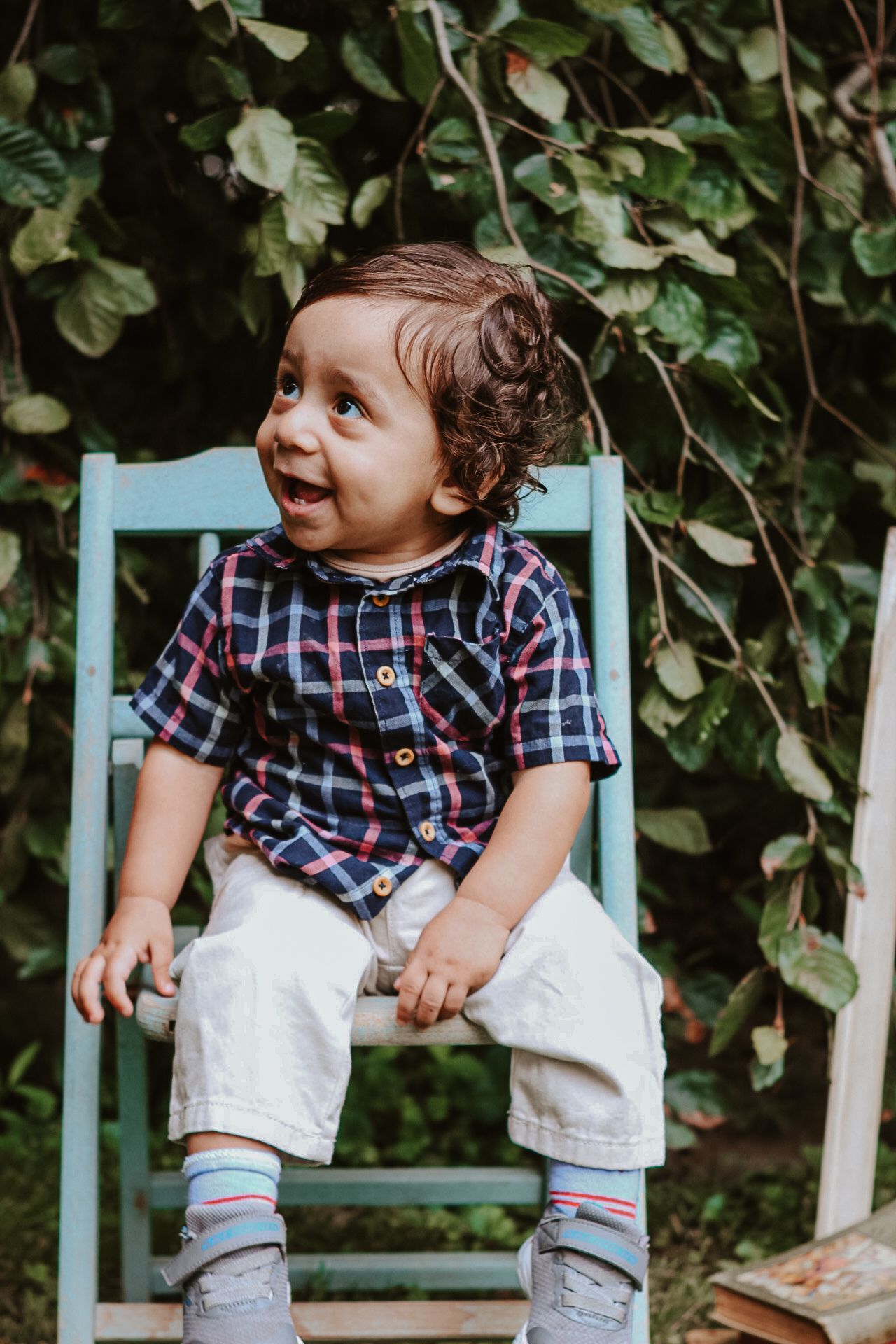Baby in plaid shirt and white pants sitting on blue chair outdoors.