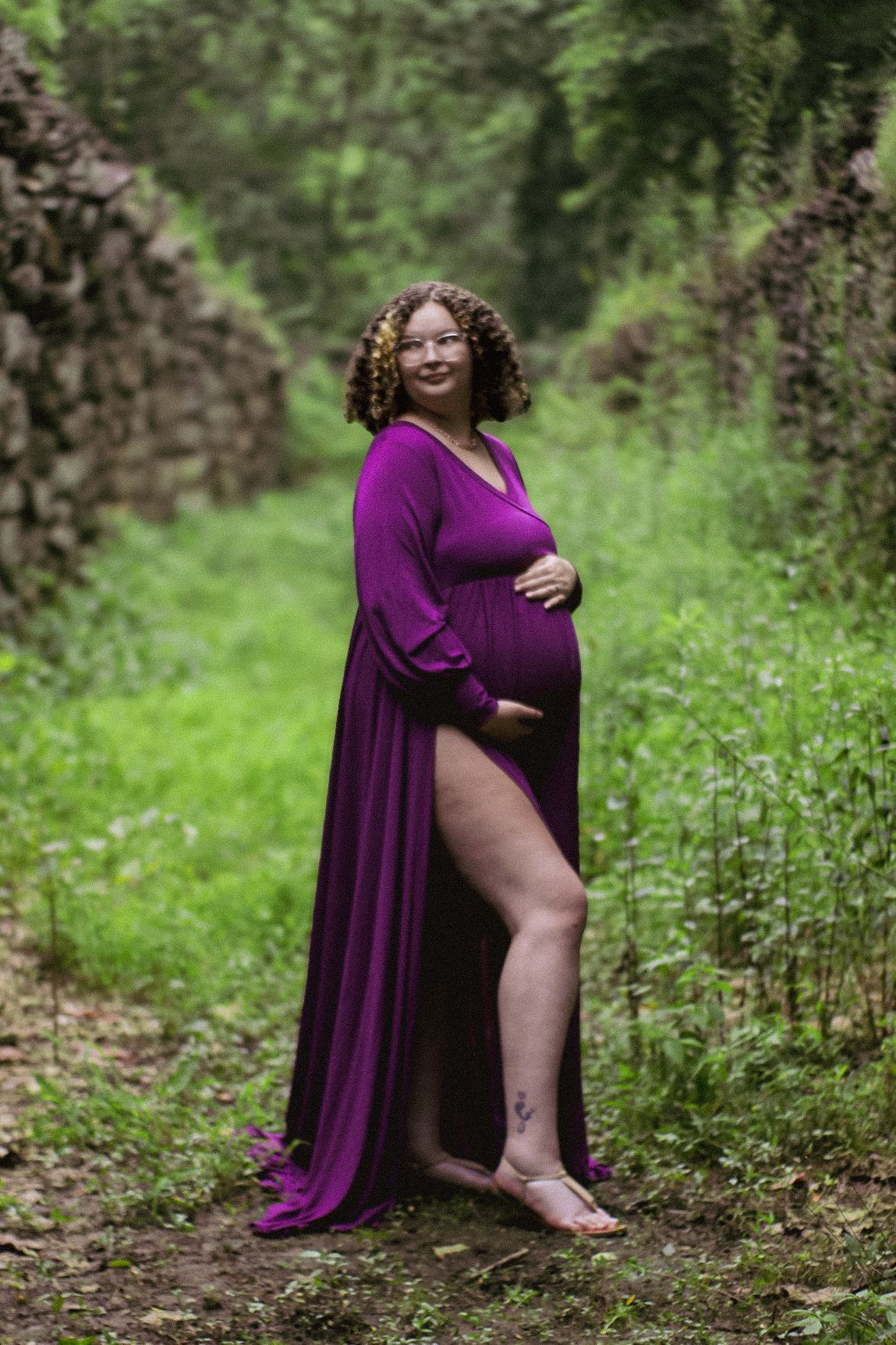 A radiant maternity portrait featuring a woman in purple dress along a stone-walled nature path.