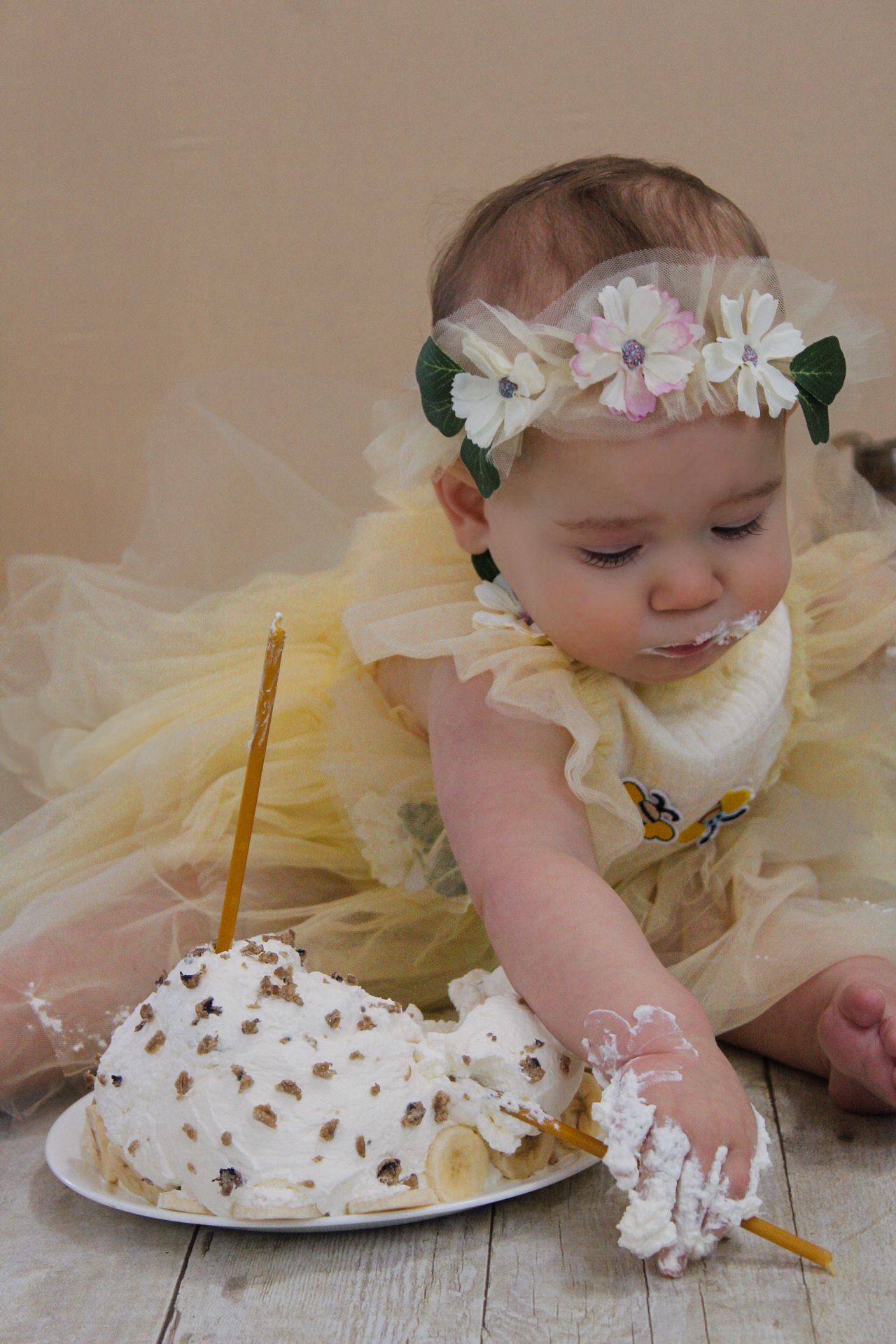 A baby in yellow dress explores a white cake with fingers on wooden floor.