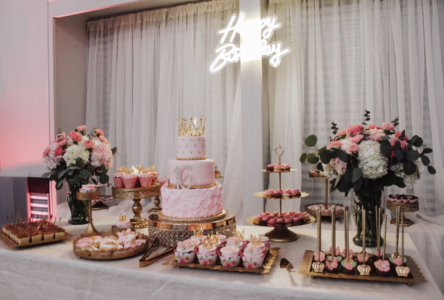 Elegant dessert table with pink flowers, tiered cake and treats under a neon sign.