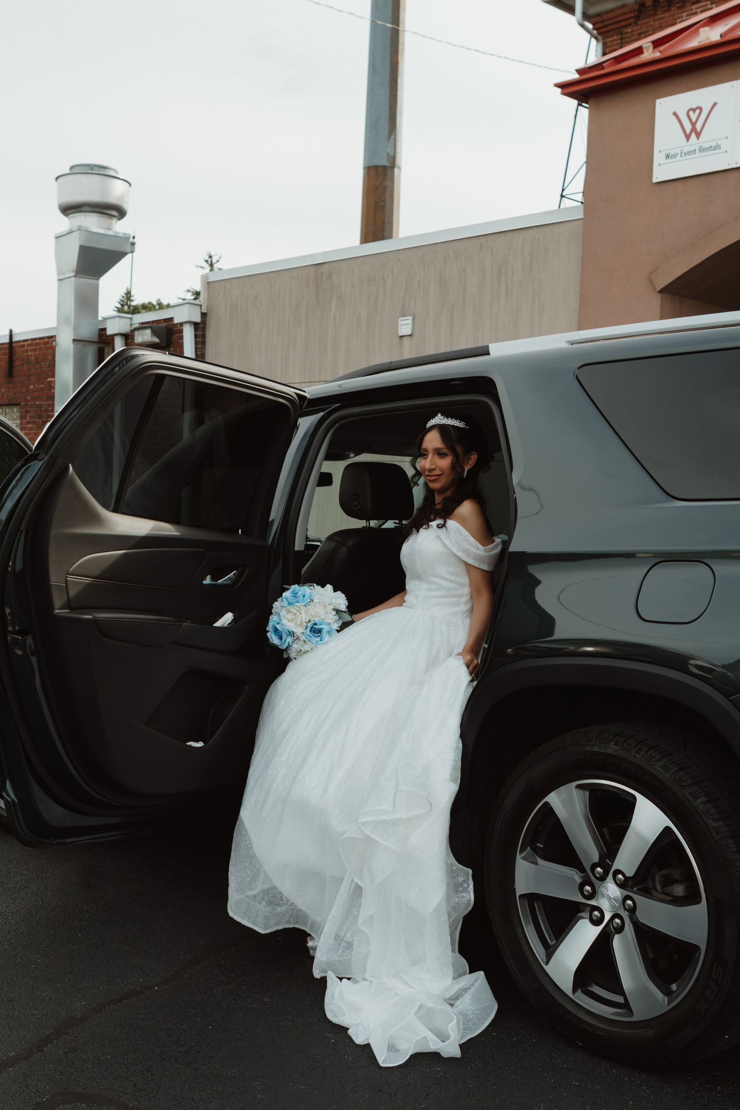 Bride in white off-shoulder gown holding blue bouquet stepping out of black SUV limo on wedding day.