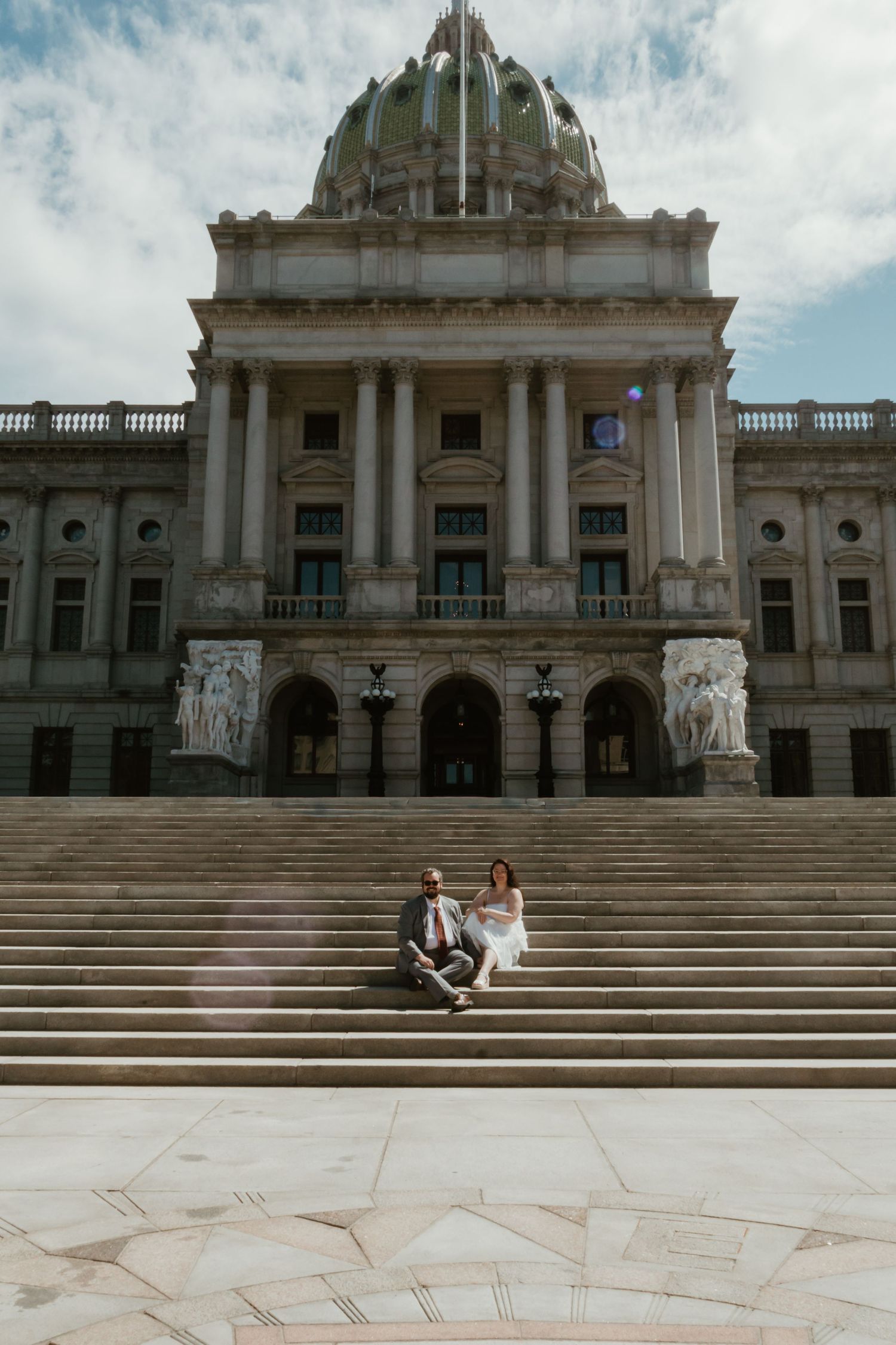 A couple sits together on the grand steps of a neoclassical capitol building under a cloudy sky.