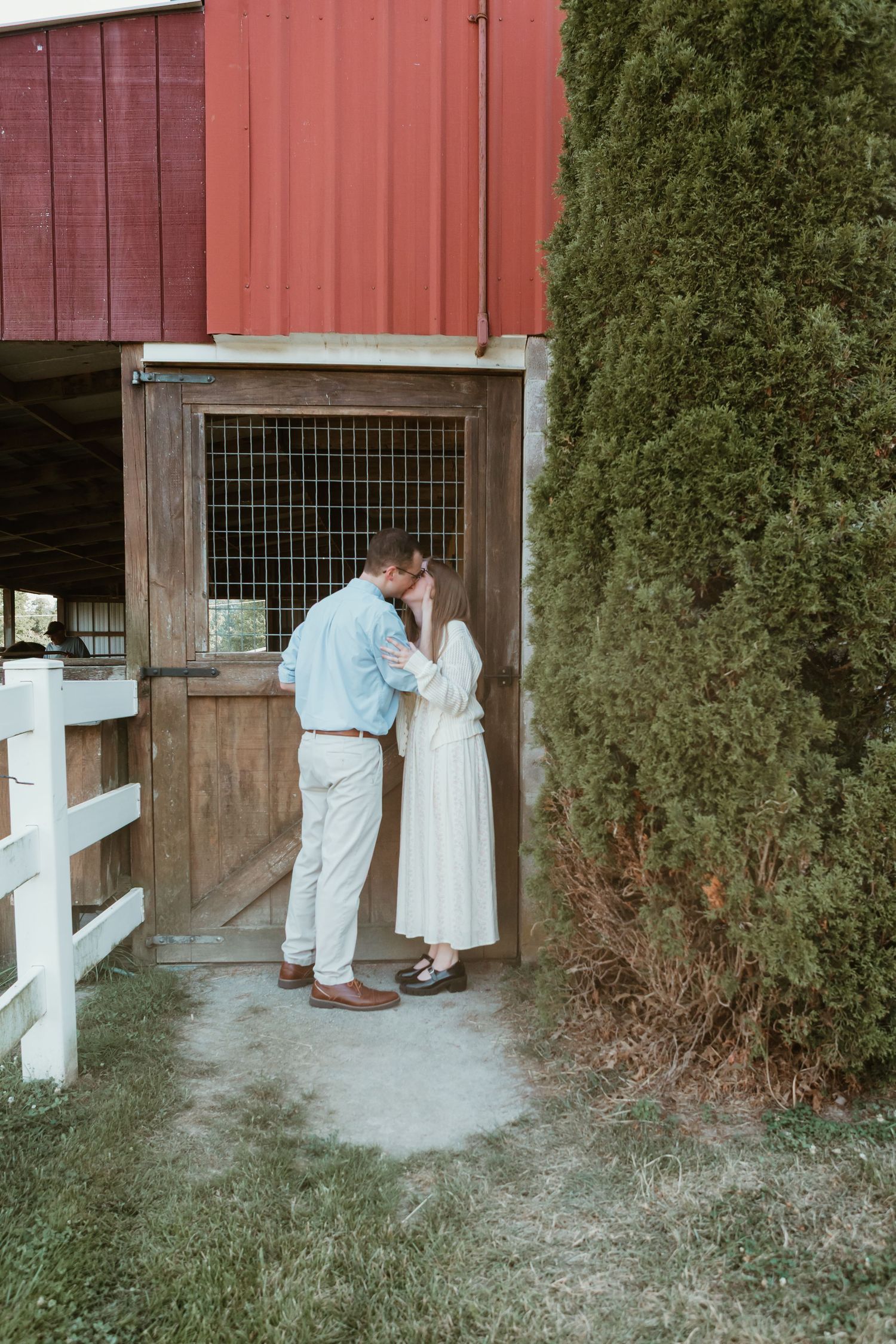 Couple sharing a romantic kiss by a rustic wooden barn door with a red barn and evergreen tree backdrop.