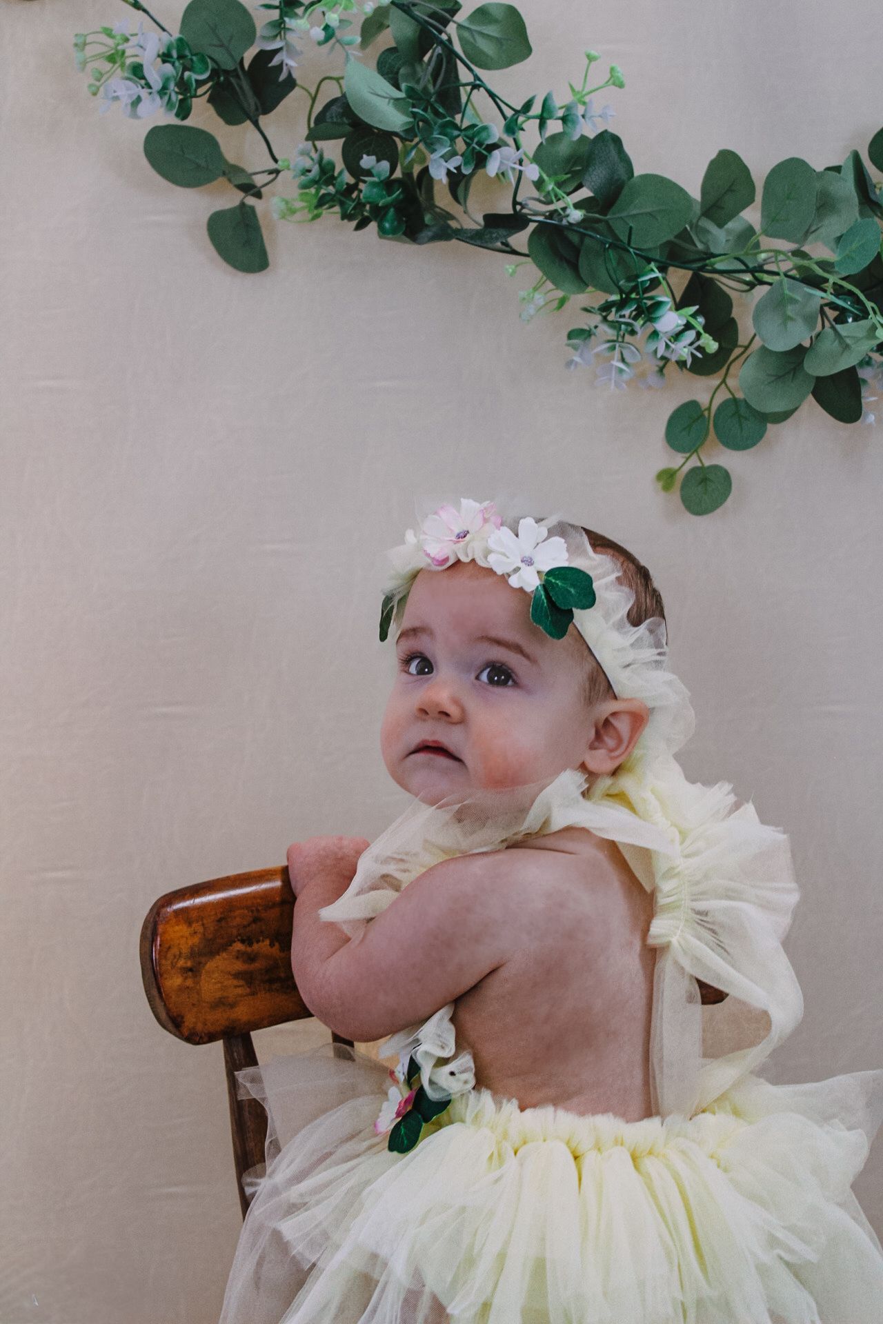 A baby in pale yellow dress sits in wooden chair under decorative greenery and flowers.