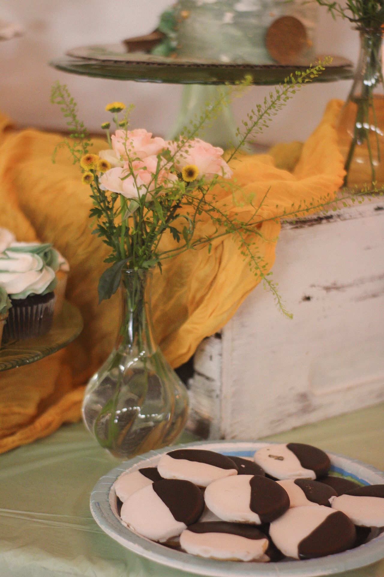 Black and pink cookies on a plate next to a vase with pink flowers and yellow fabric.