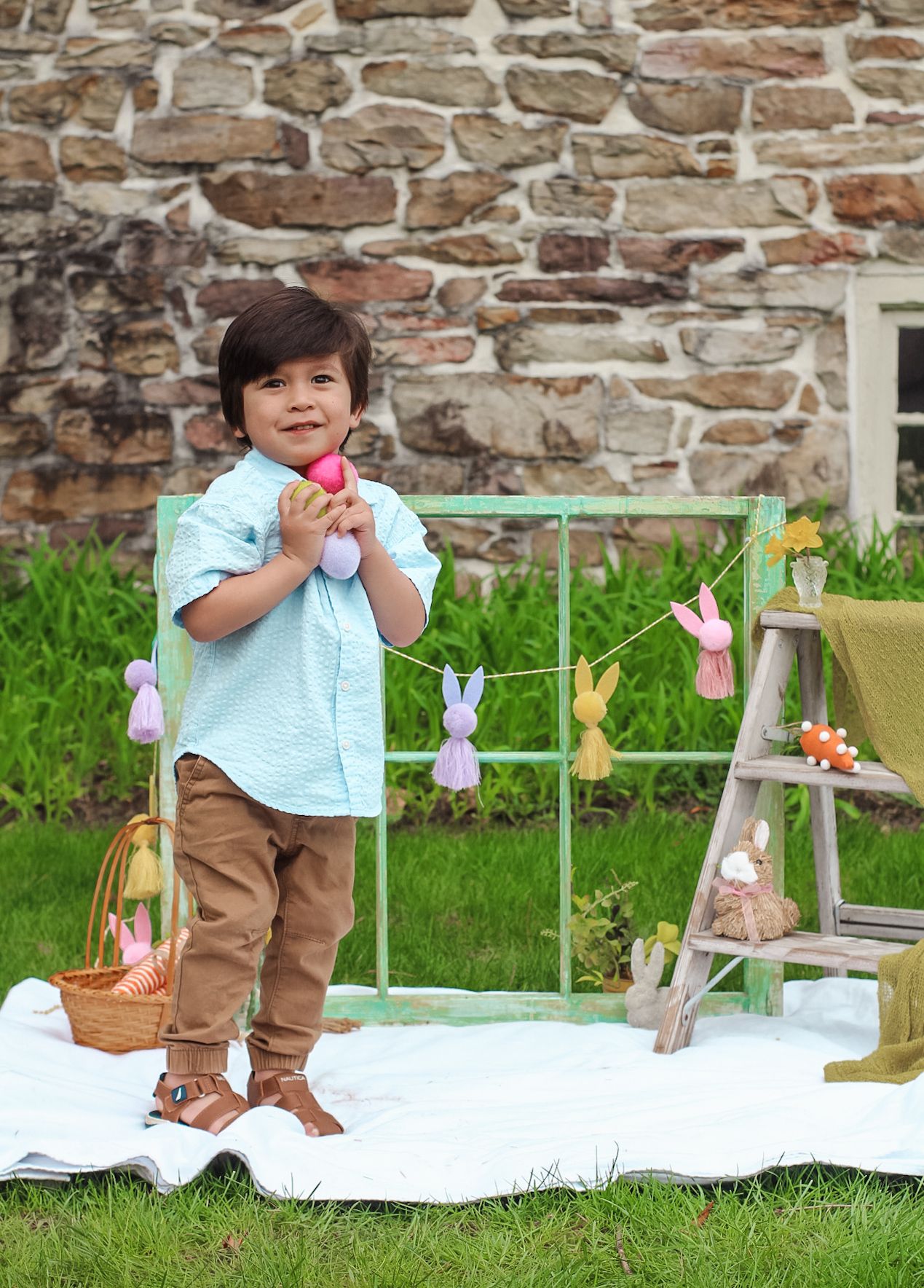 Easter picnic setup outdoors with decorative eggs, bunnies, and spring decor on white blanket against stone wall.