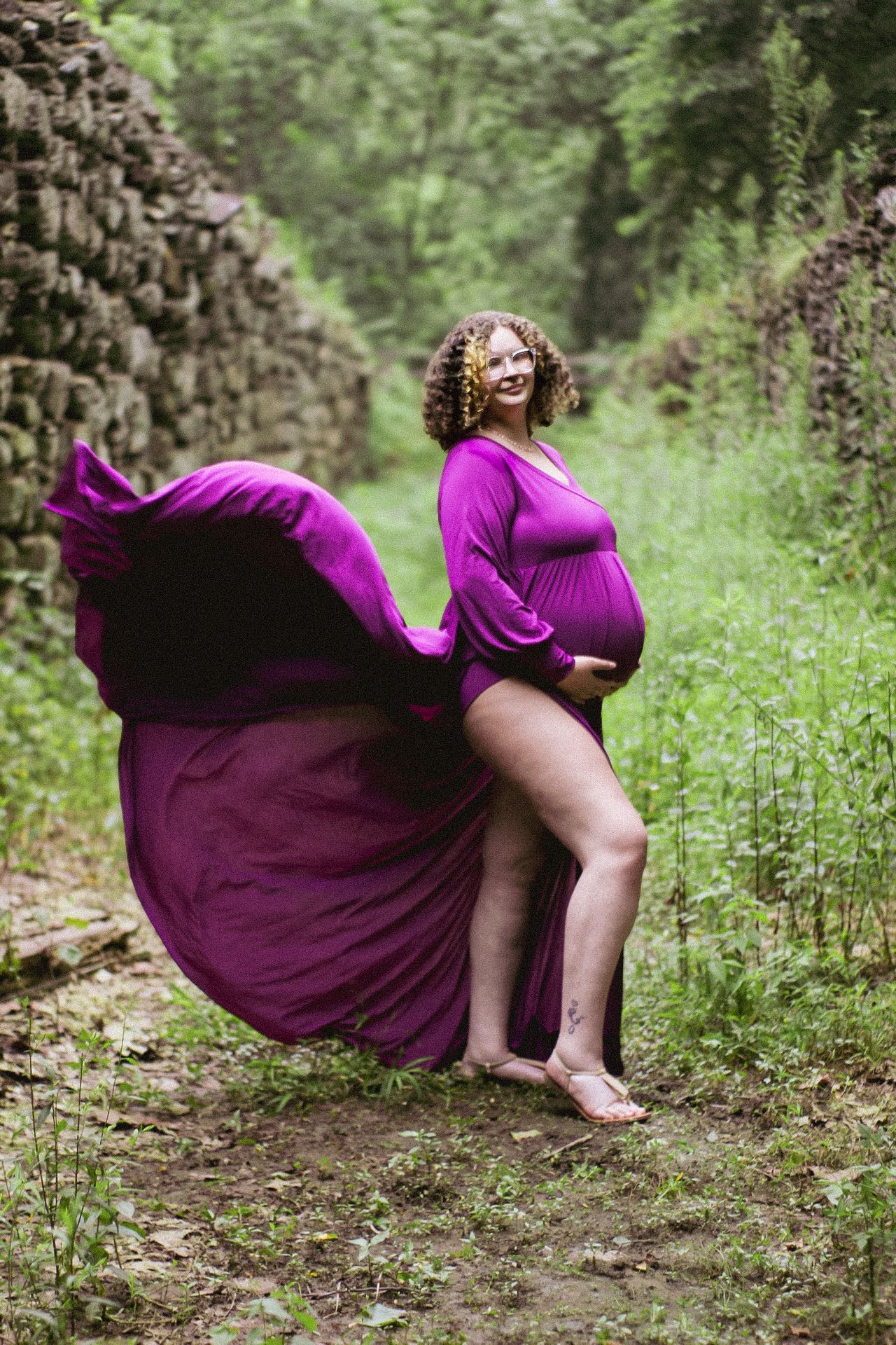 A pregnant woman in a flowing purple dress poses dramatically in a wooded path with stone walls.