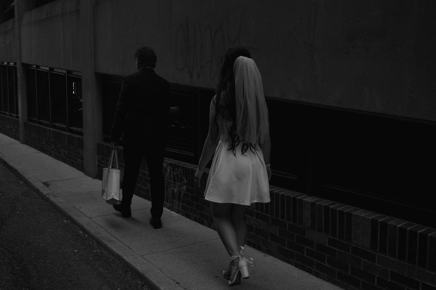 Black and white photo of a bride in white dress and veil walking with a man in suit on a city sidewalk.