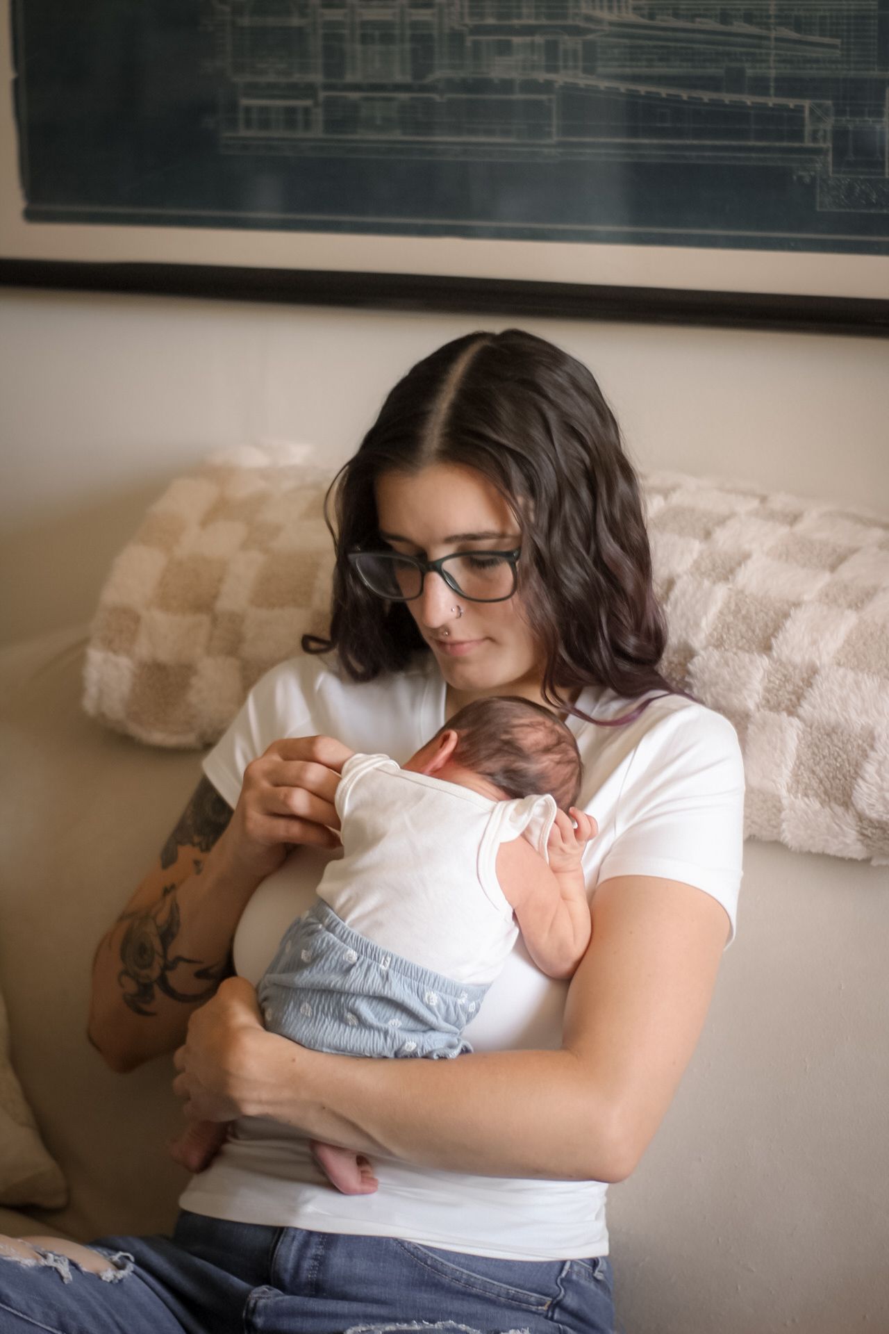 Parent in glasses and white t-shirt holds sleeping infant on beige couch.