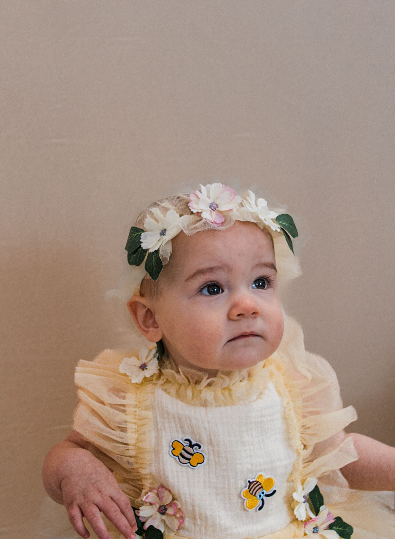 A baby wearing a yellow dress with bee decorations and a floral crown looks up with curiosity.
